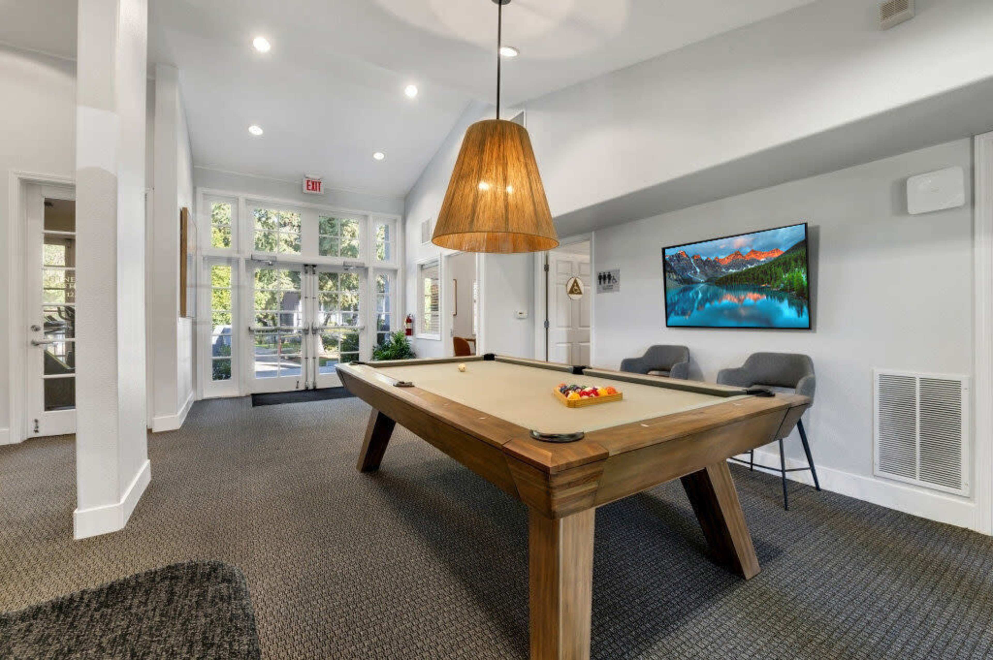 The image shows a bright, modern game room featuring a billiards table, a large pendant light, and a flat-screen TV on the wall, with large windows providing ample natural light.