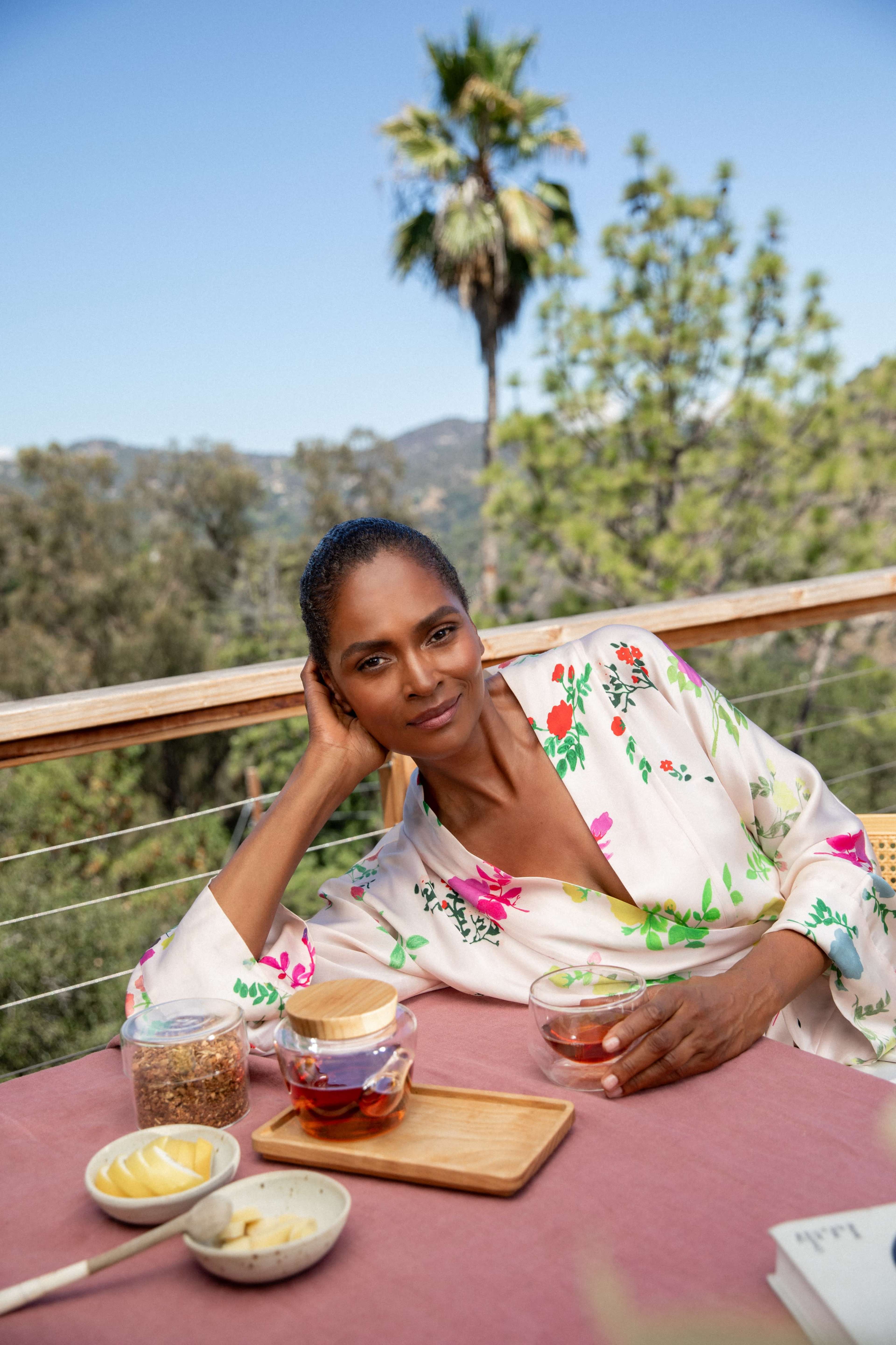 A woman in a floral robe is seated at a table on a balcony, holding a glass of tea, with a scenic view of trees and mountains in the background.