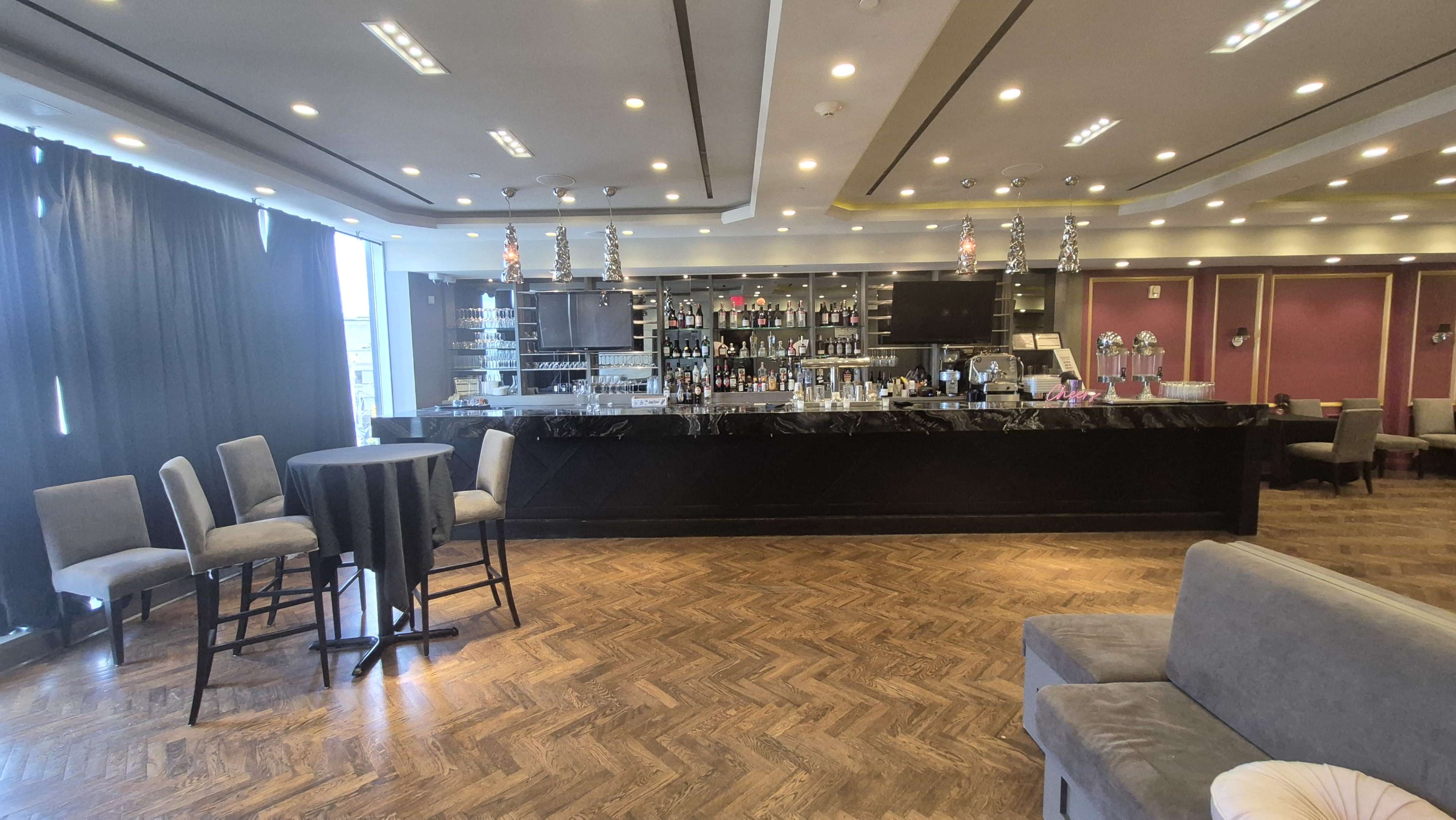 The image shows a modern bar area with a large countertop, shelves filled with various bottles, and seating arrangements including high stools and a round table.