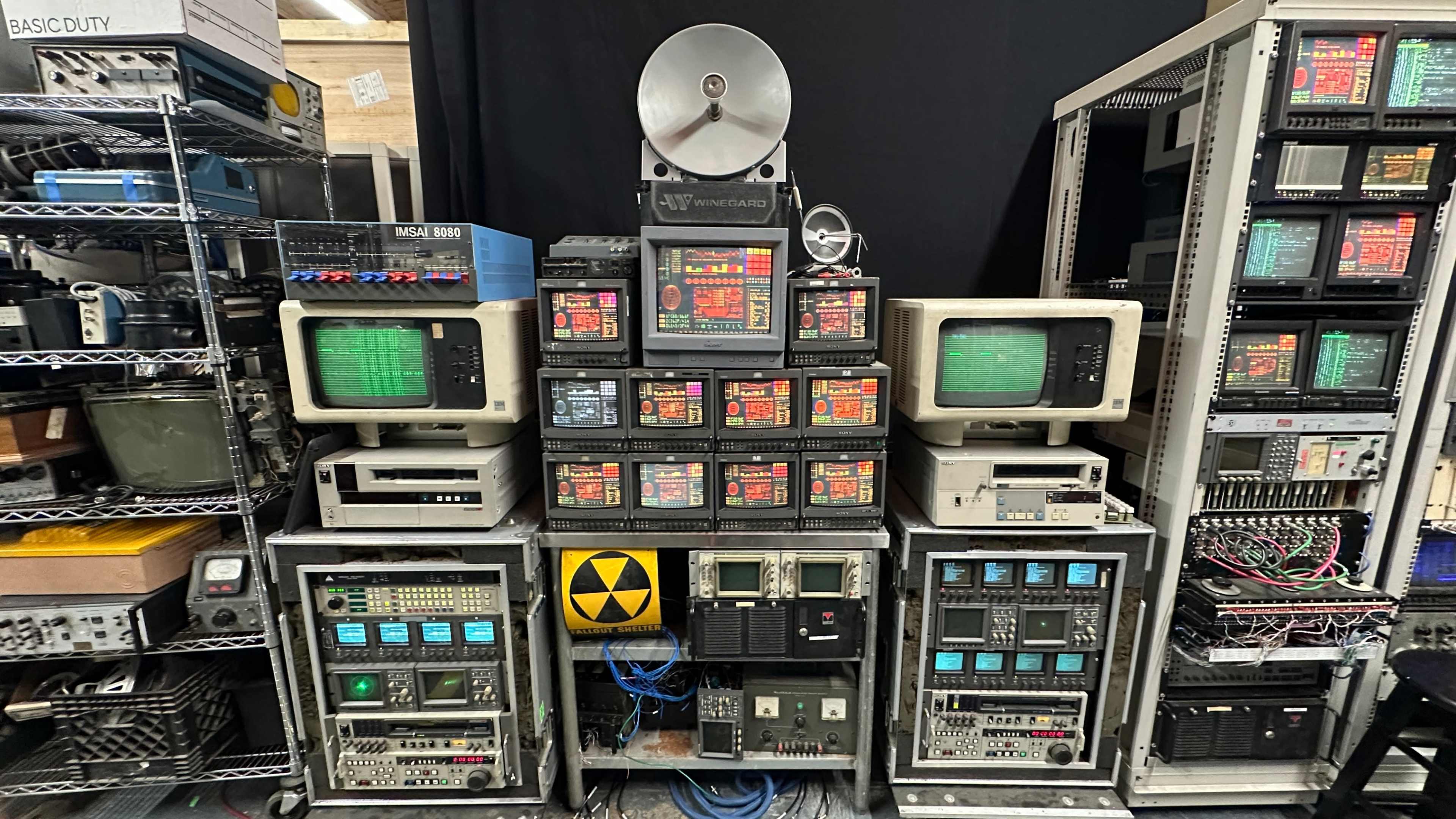 The image shows an arrangement of vintage electronic equipment, including multiple monitors, control panels, and machinery, set against a black backdrop.