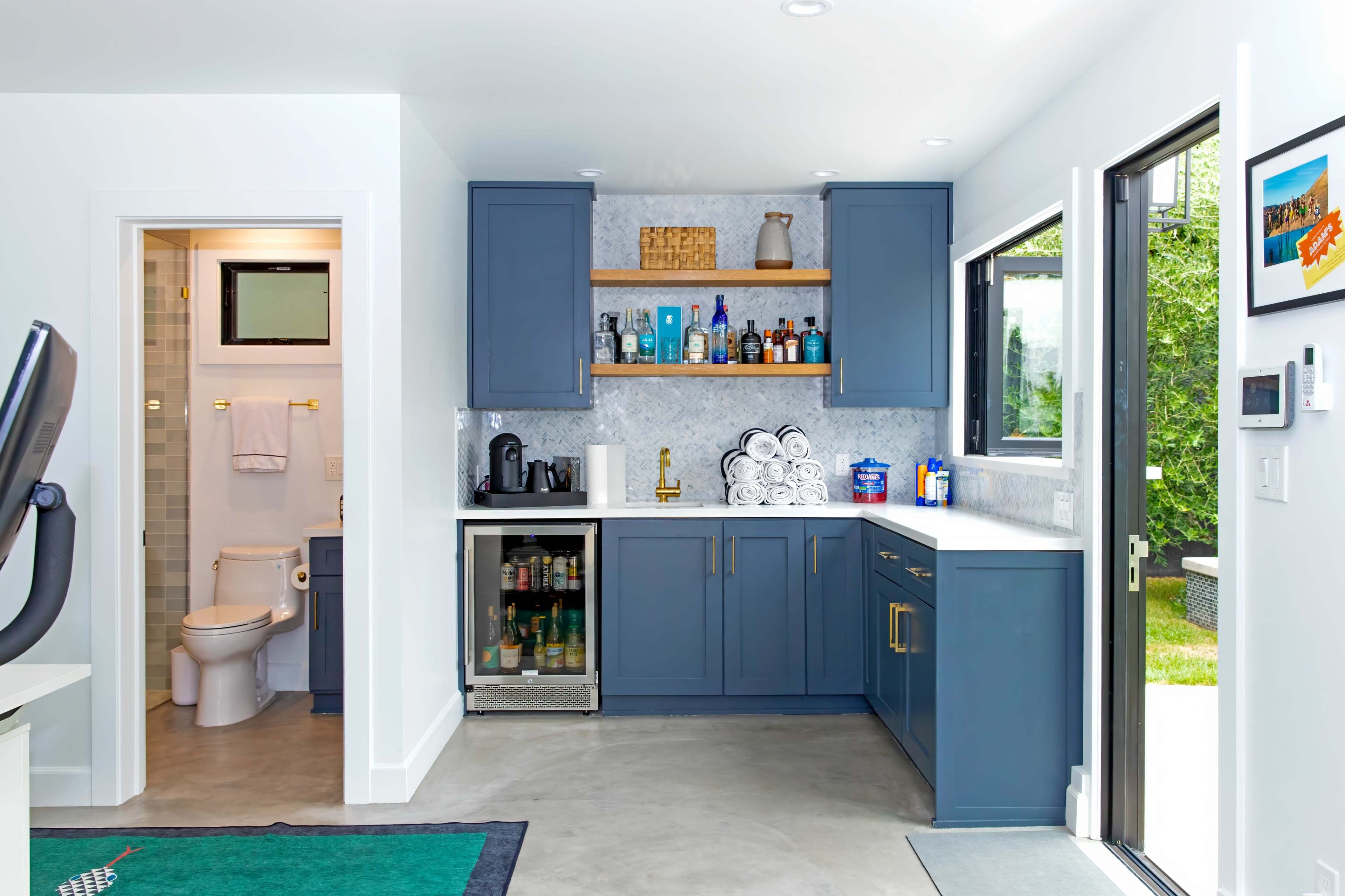 A modern, organized kitchen space with blue cabinetry, a countertop, and a small beverage fridge, adjacent to a bathroom area.