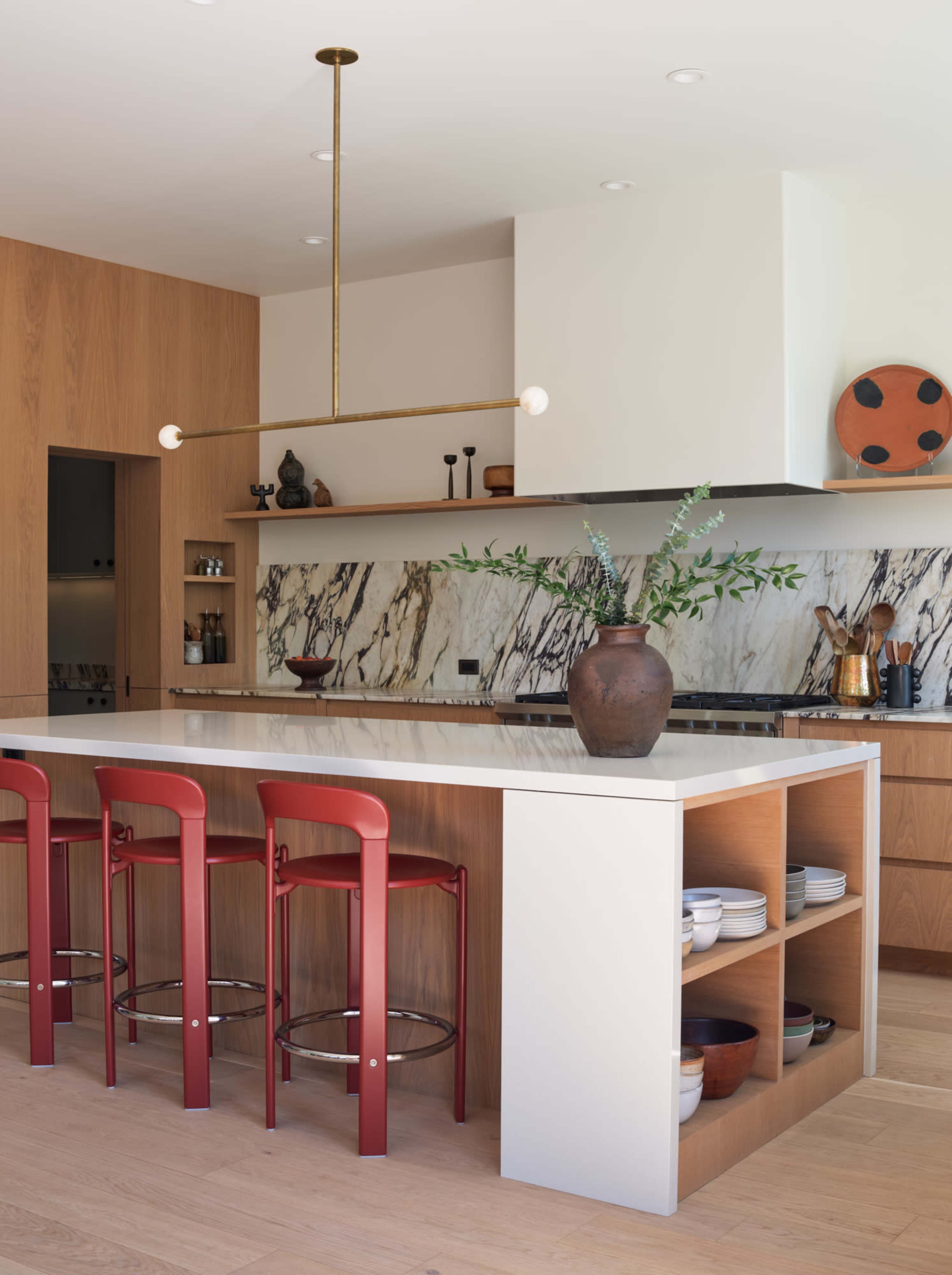 The image depicts a modern kitchen with a long island featuring four red bar stools, a marble backsplash, and various kitchen items staged on the shelves.