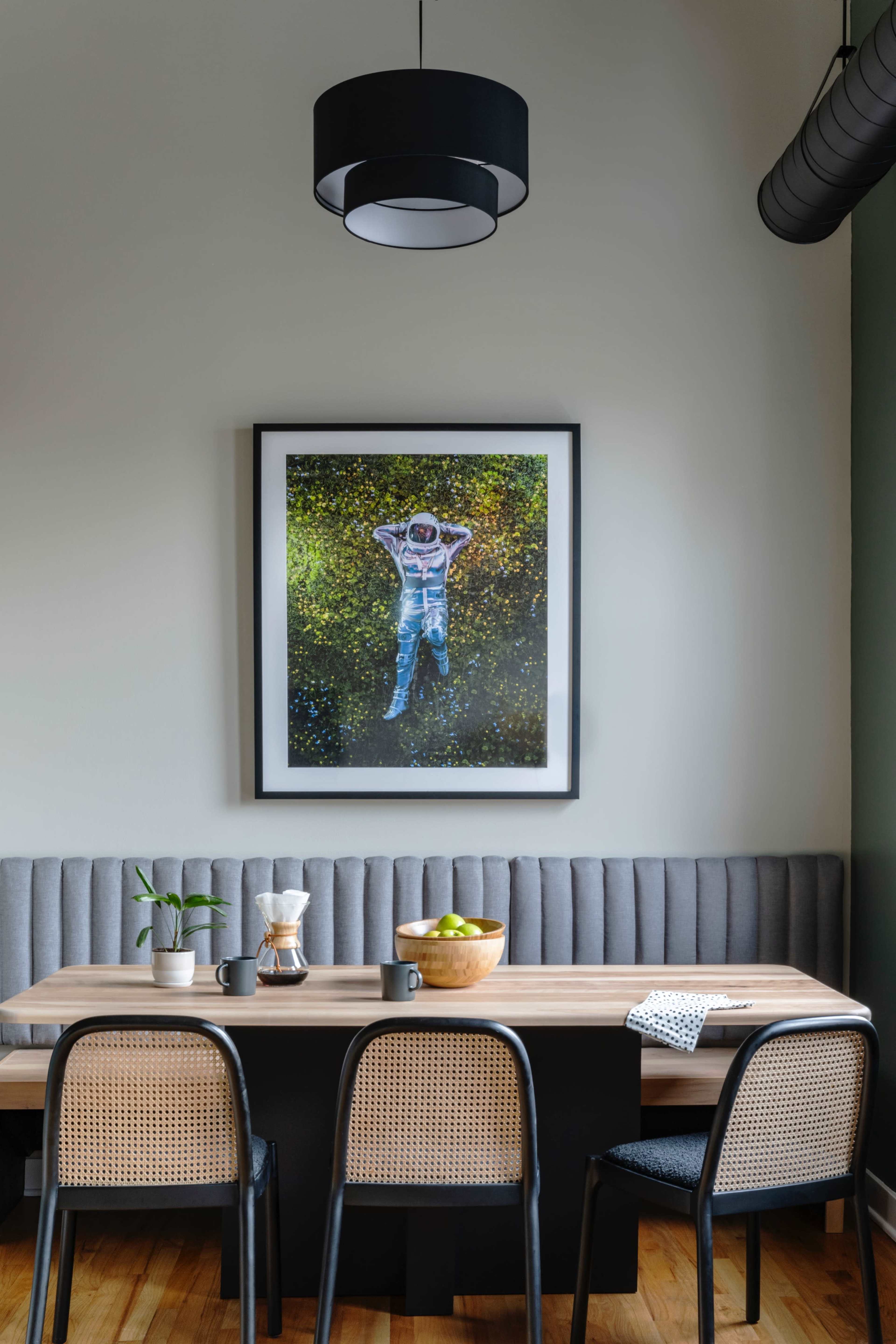 A dining area features a wooden table surrounded by black and cane chairs, with a framed astronaut artwork hanging on the wall above a gray upholstered bench.