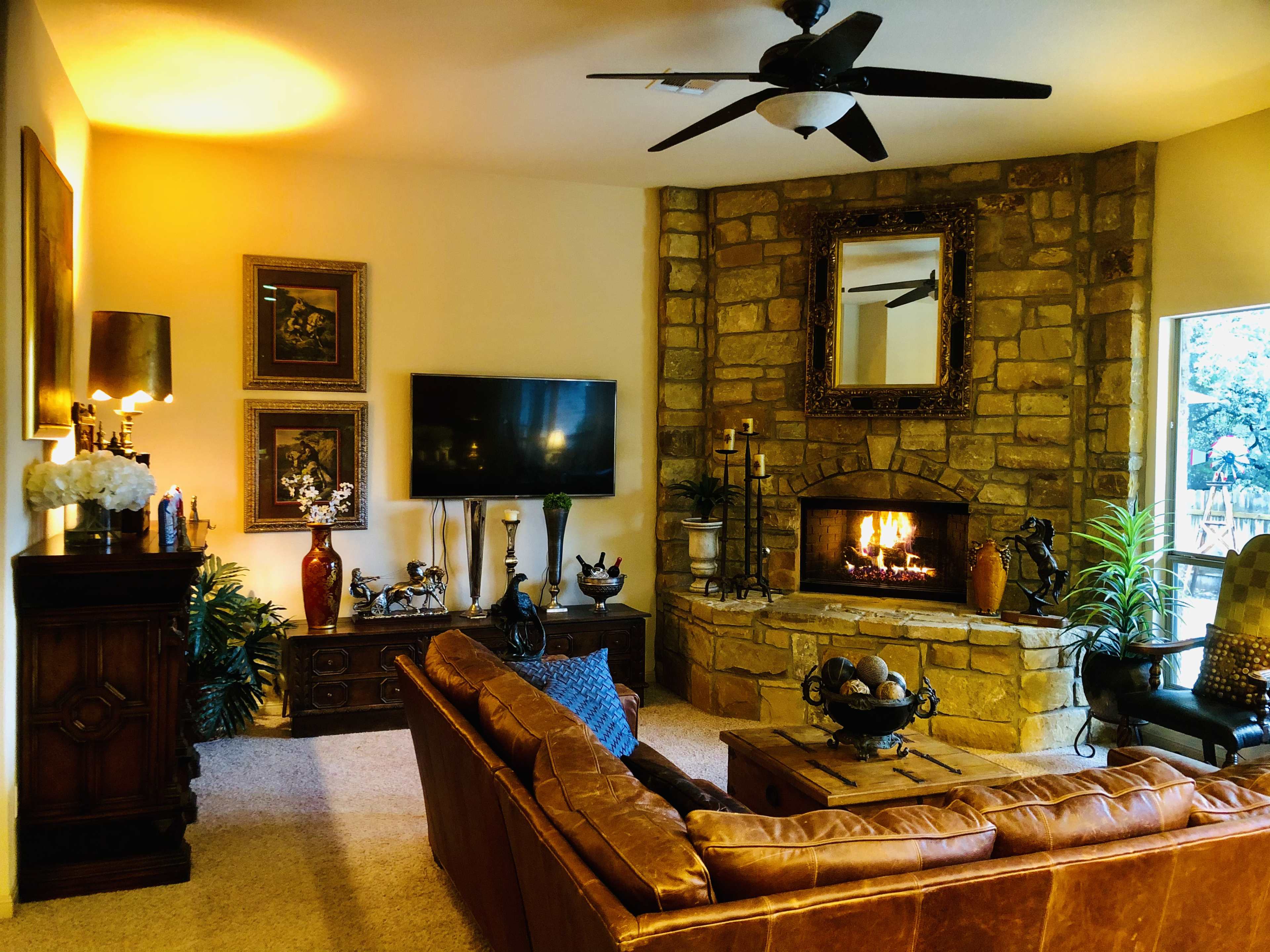 A warmly decorated living room with a stone fireplace, a television mounted on the wall, and a leather sofa arranged around a coffee table.