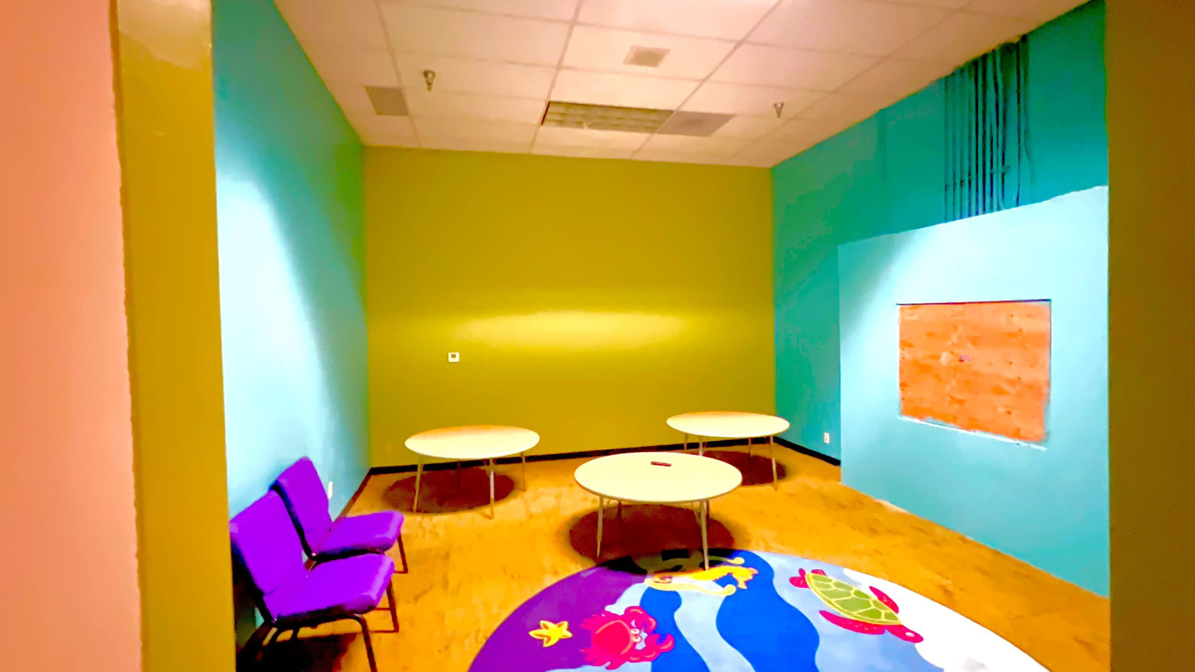 The image shows a brightly colored room featuring four round tables, a small purple chair, and a carpet with a design.