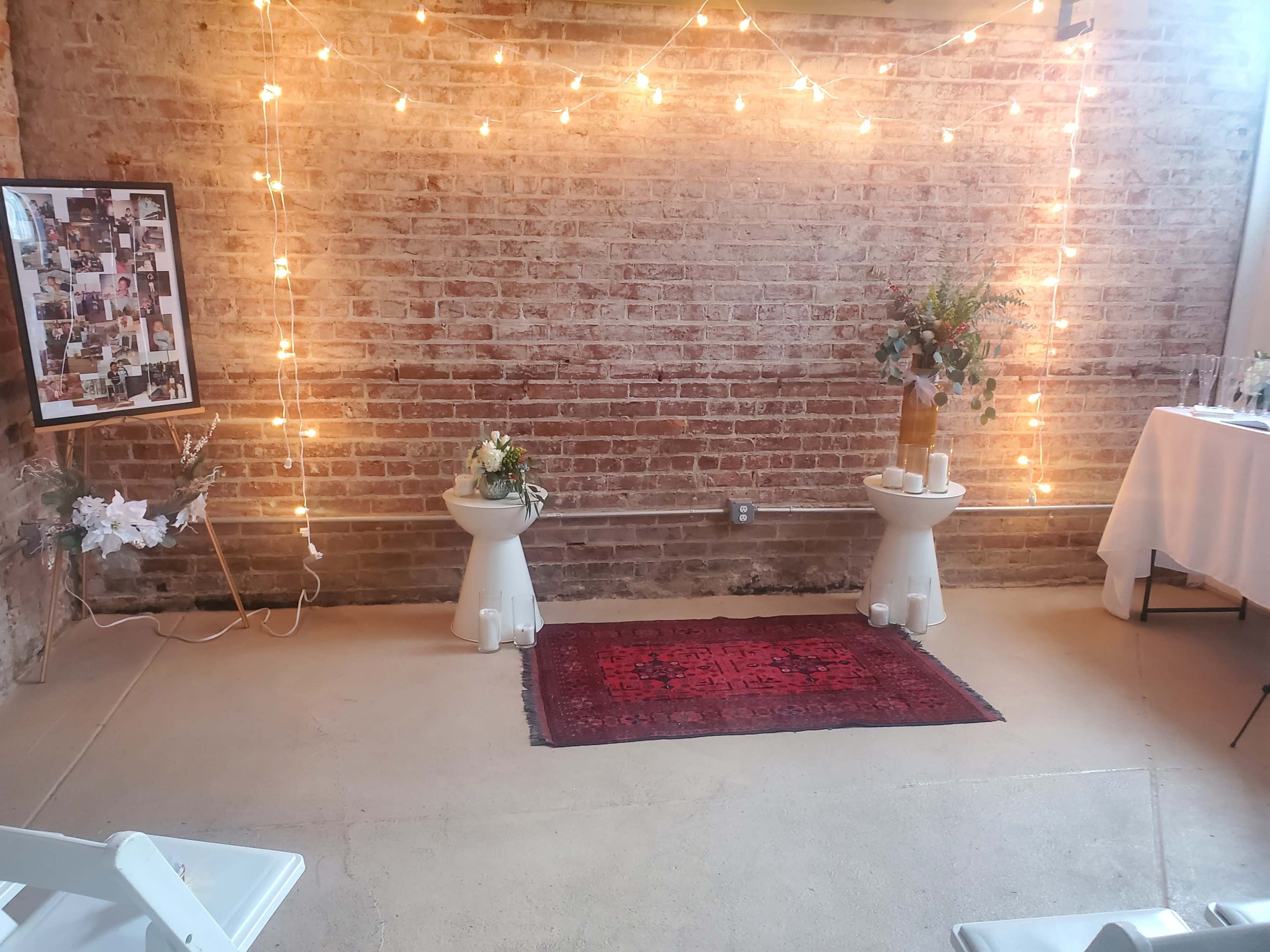 A rustic interior space with brick walls, string lights hanging above, two small white pedestals with floral arrangements, and a red area rug on the floor.
