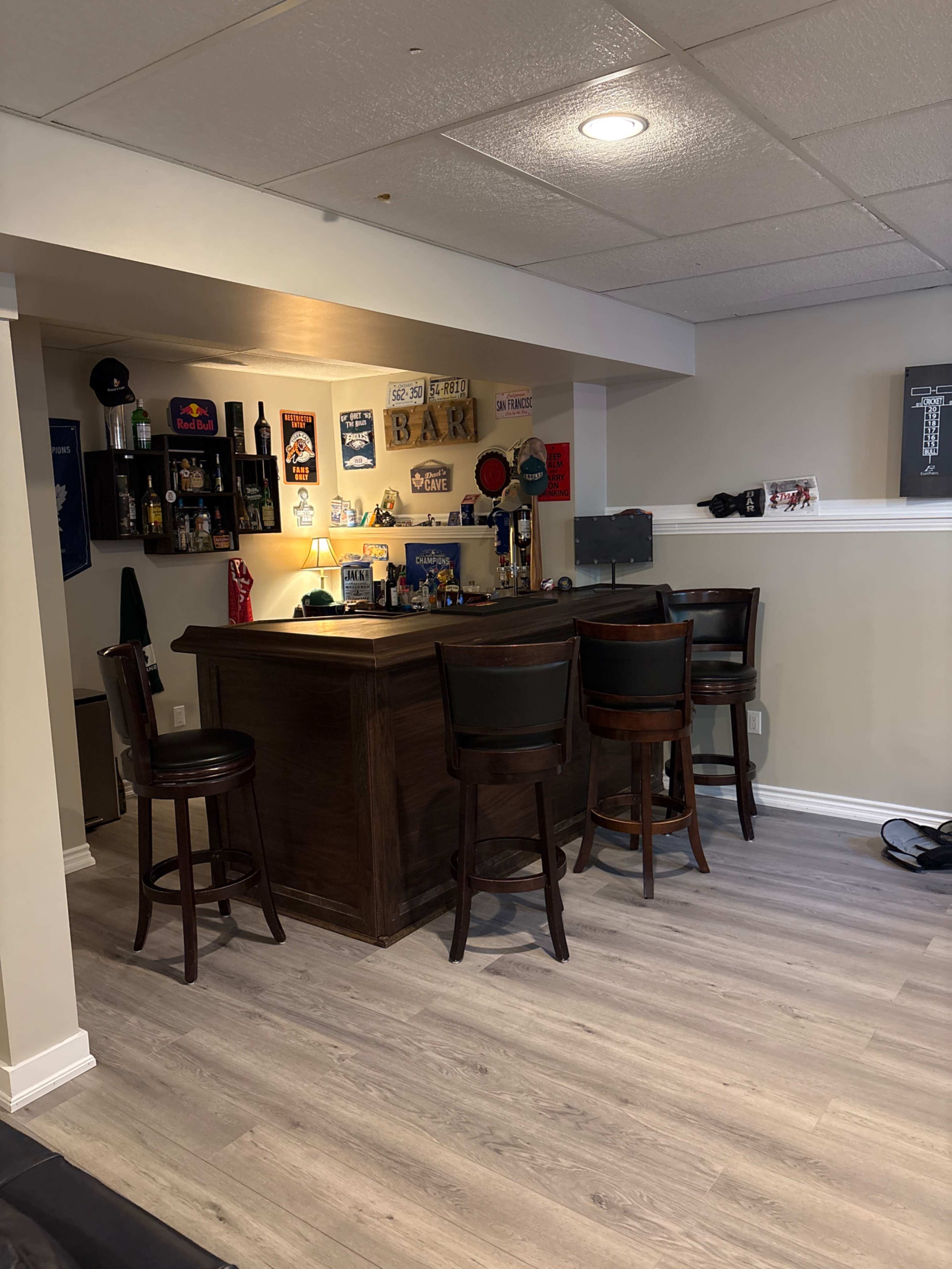 The image shows a home bar area with four wooden stools around a dark wooden bar, decorated with various bottles and memorabilia on the walls.