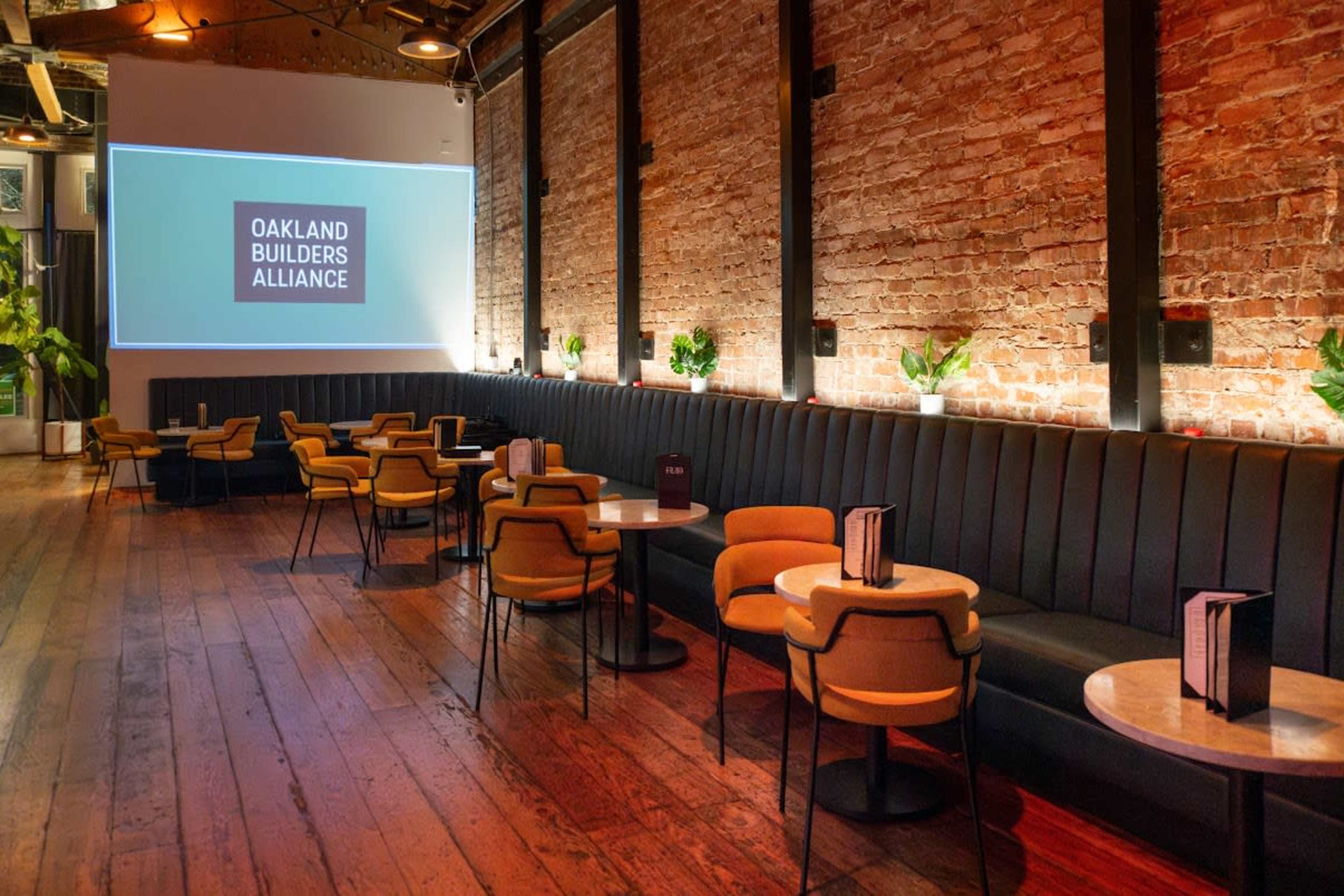 The image shows a spacious venue with a brick wall, wooden flooring, and seating that includes circular tables and yellow chairs, featuring a screen displaying "Oakland Builders Alliance."