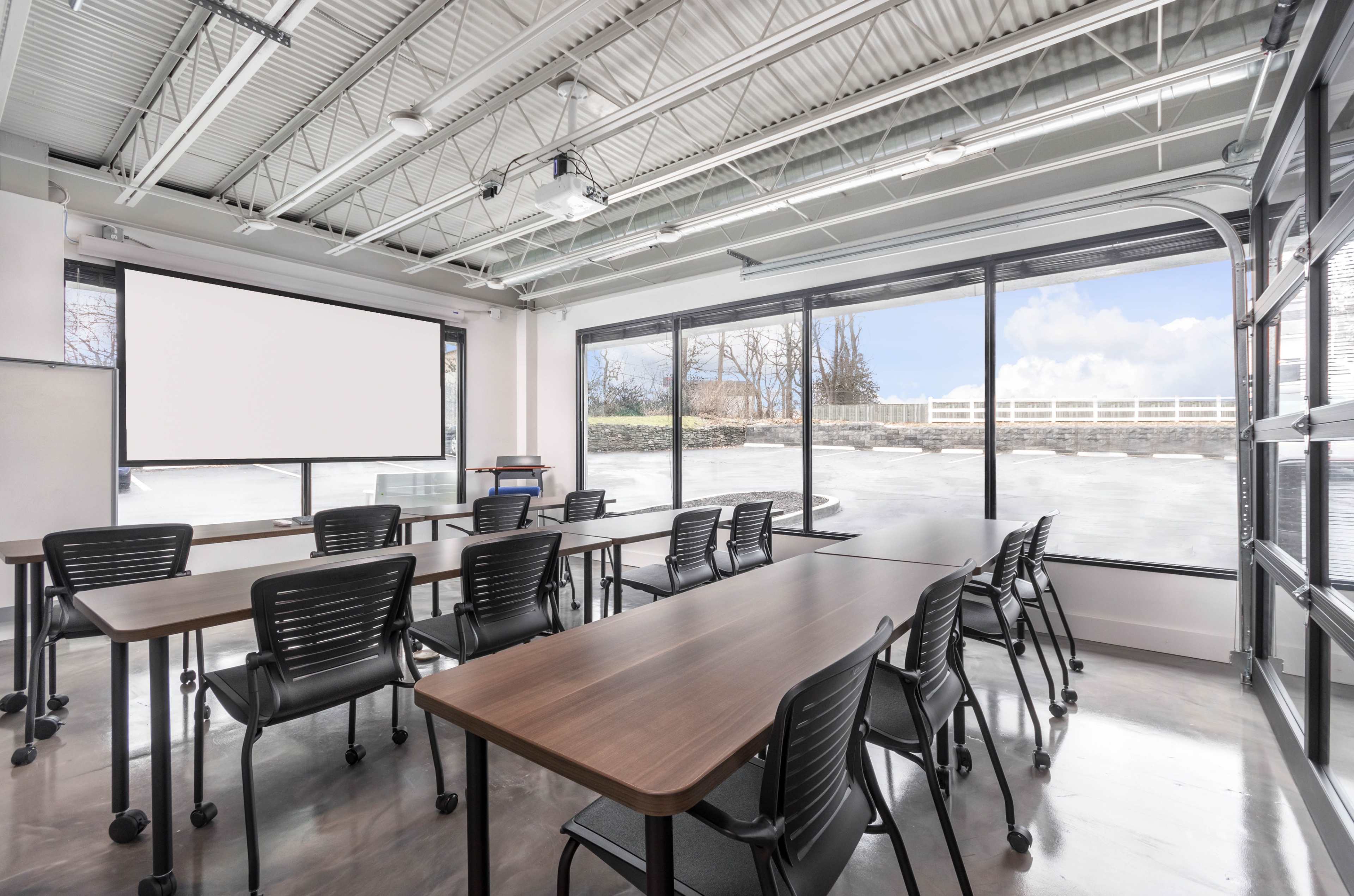 A modern classroom features multiple tables arranged in a U-shape, with black chairs, and large windows overlooking an outdoor area.