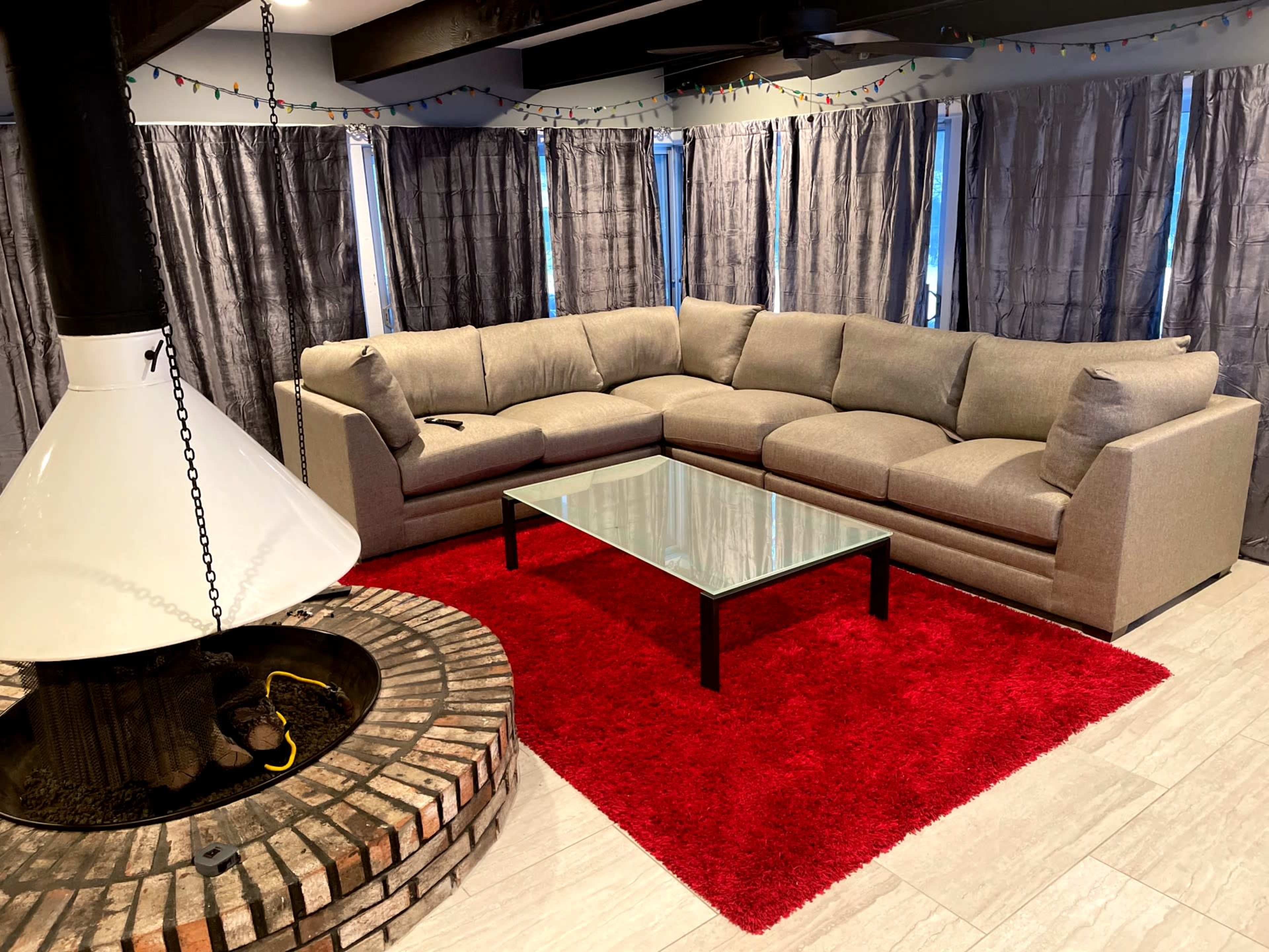 The image shows a modern living room featuring a large sectional sofa, a glass coffee table, and a circular brick fireplace on a red area rug.