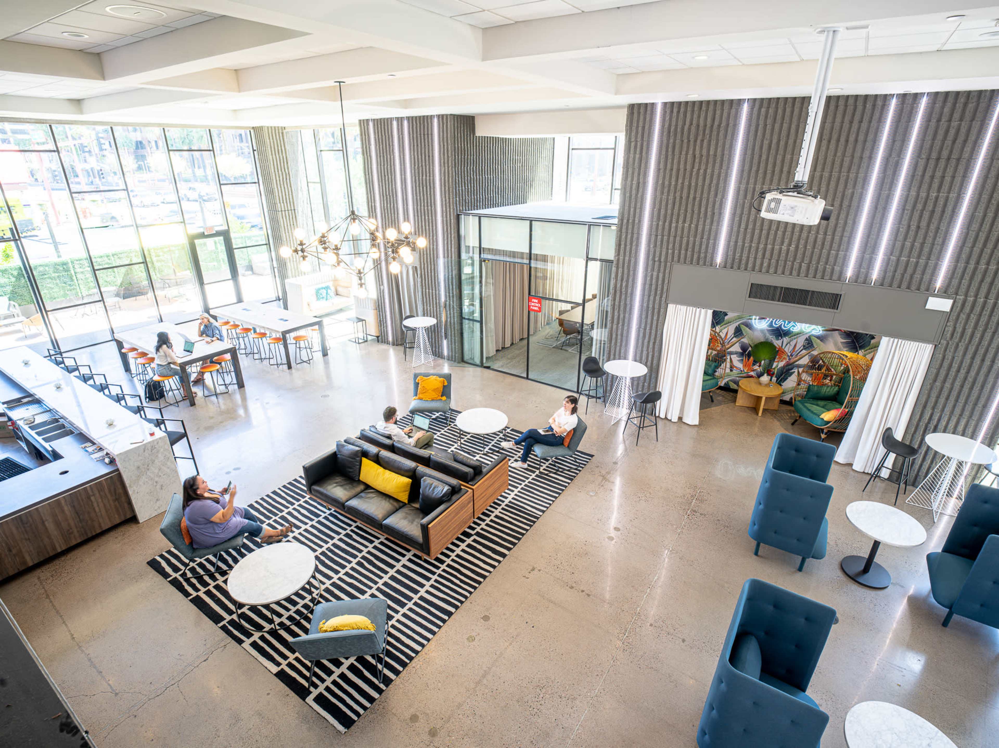 The image shows a modern, open lounge area featuring a sectional sofa, several small tables, and a large window allowing natural light to enter.