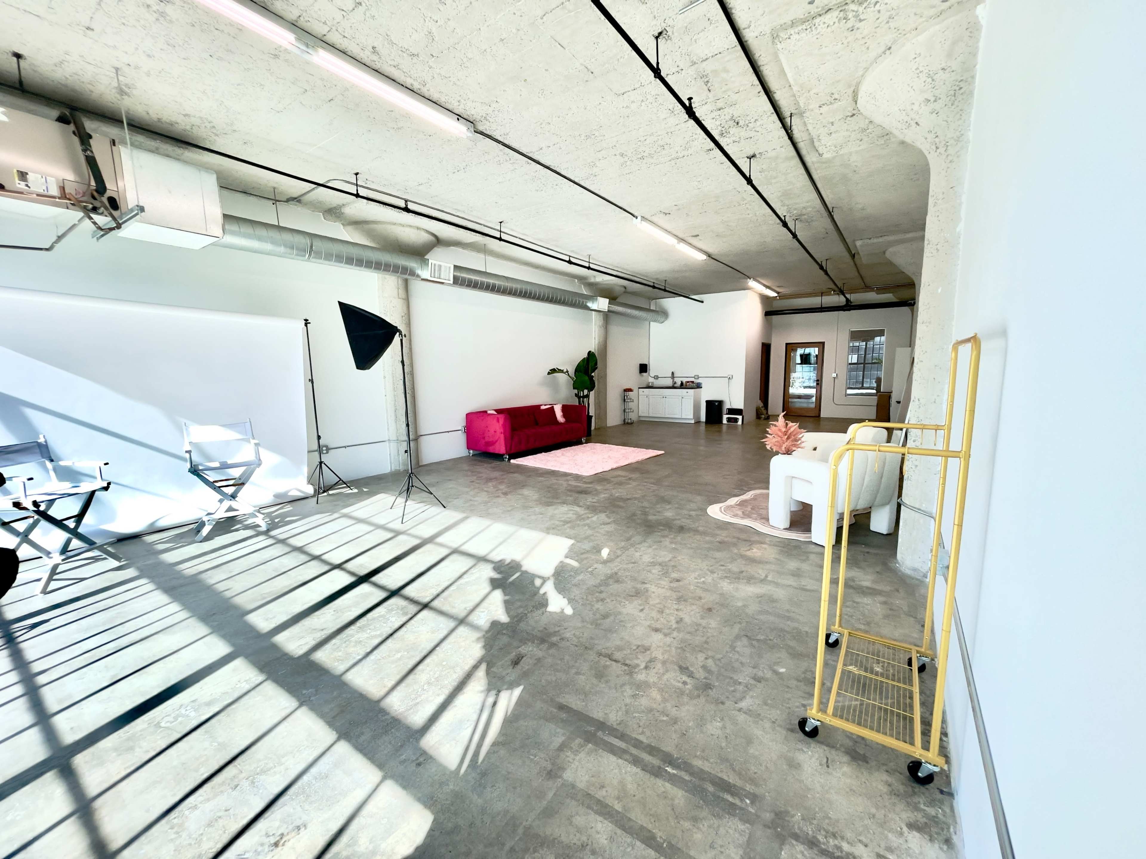 The image shows a spacious, minimalist studio interior with concrete flooring, a pink sofa, a small kitchenette, and a large window allowing natural light to fill the room.