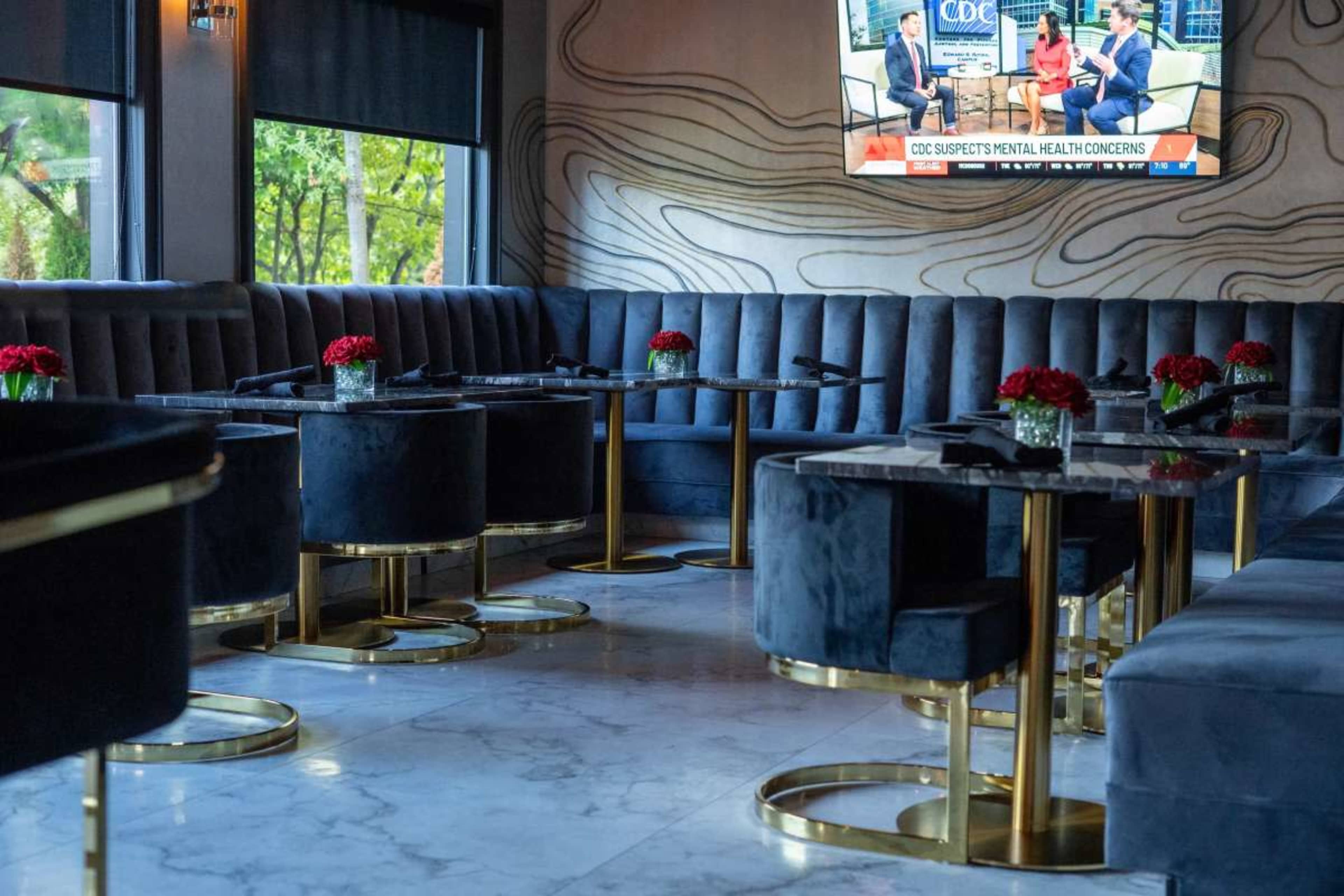 The image shows a modern, upscale restaurant interior with plush dark seating, elegant circular tables, and a television displaying a news segment in the background.