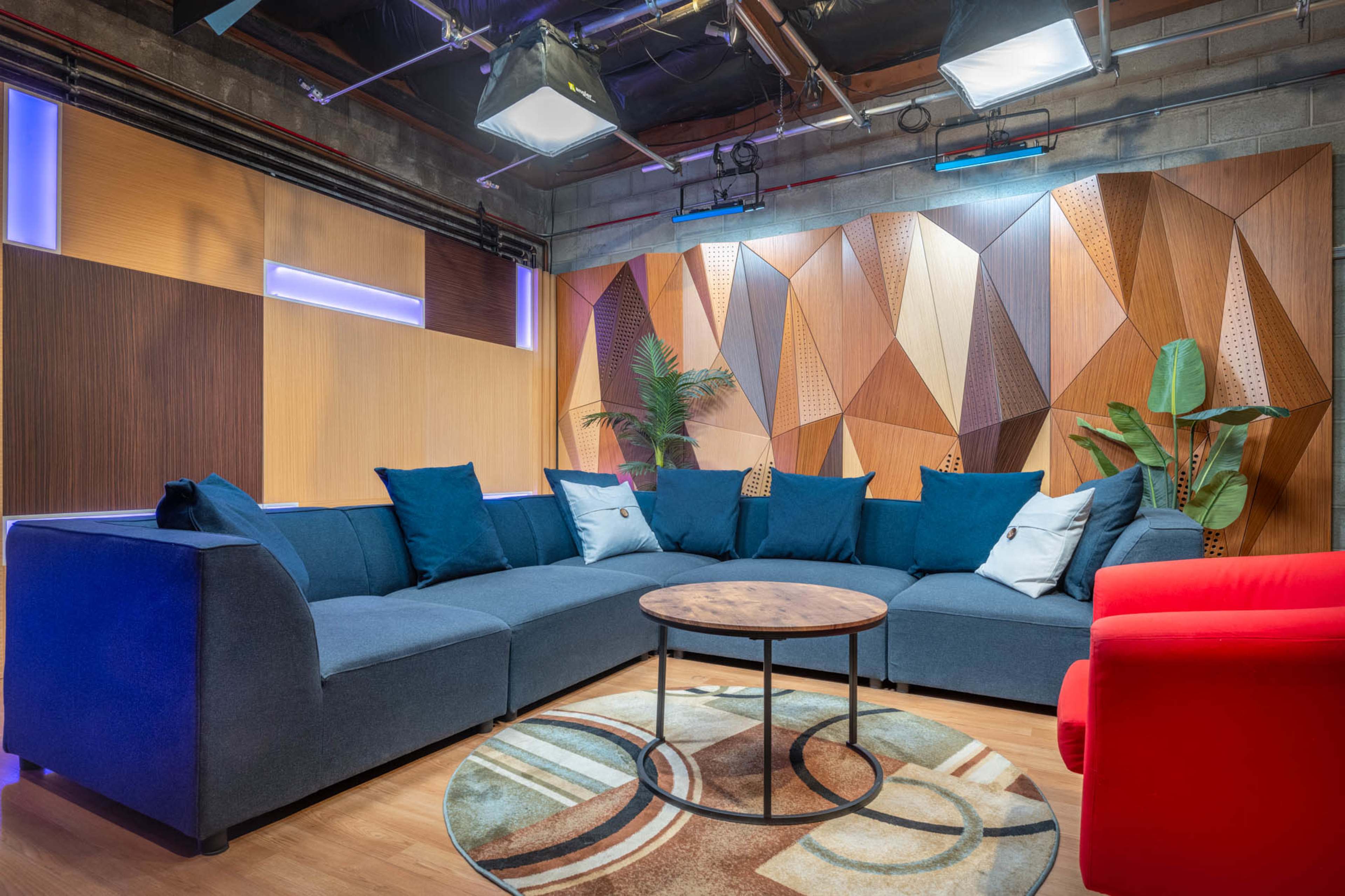A modern lounge area featuring a large sectional sofa with blue cushions, a round wooden coffee table, and a red accent chair, all set against a geometric wall design and indoor plants.