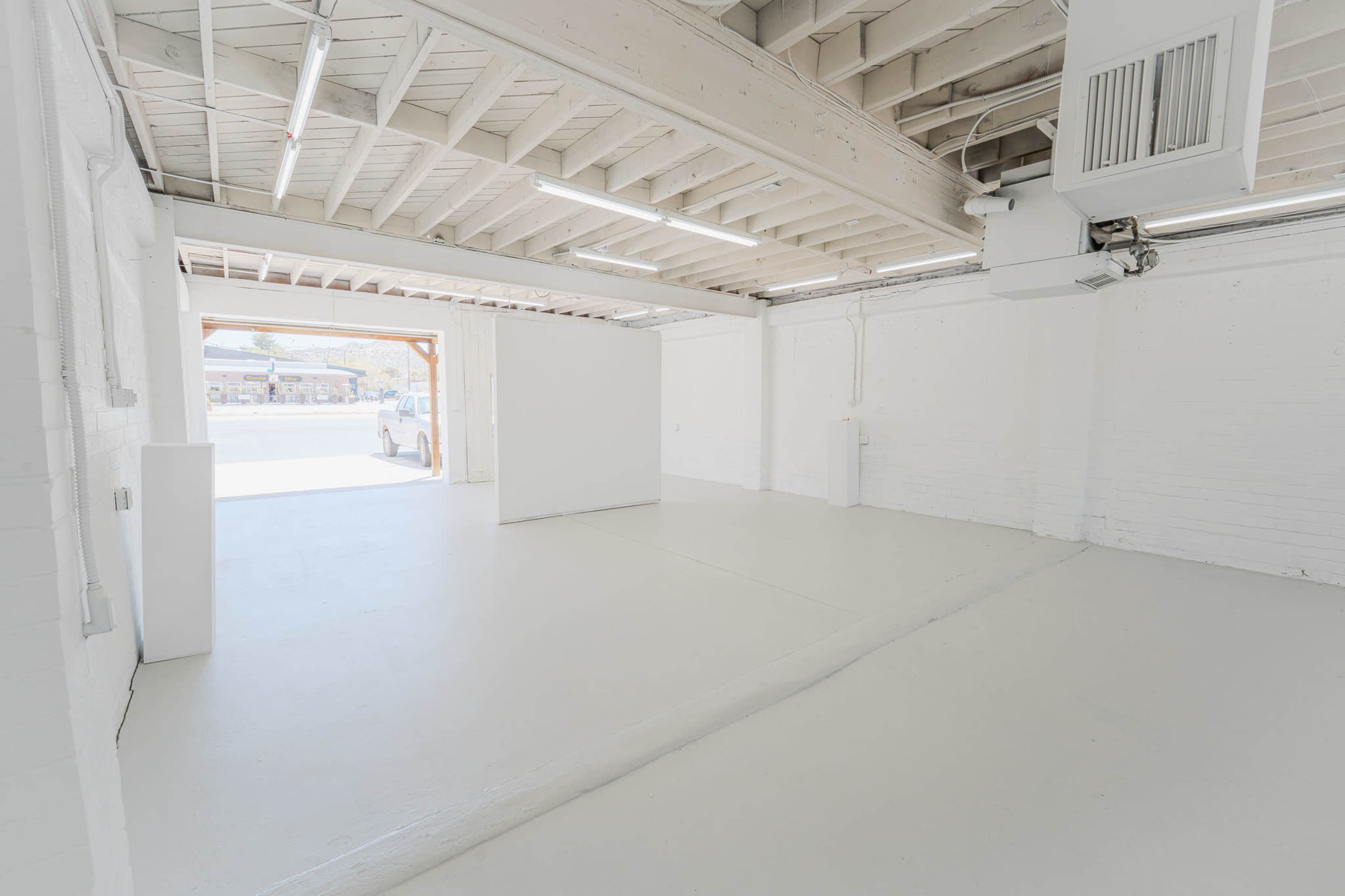 An empty white interior space with a concrete floor, exposed wooden beams, and a doorway leading outside.
