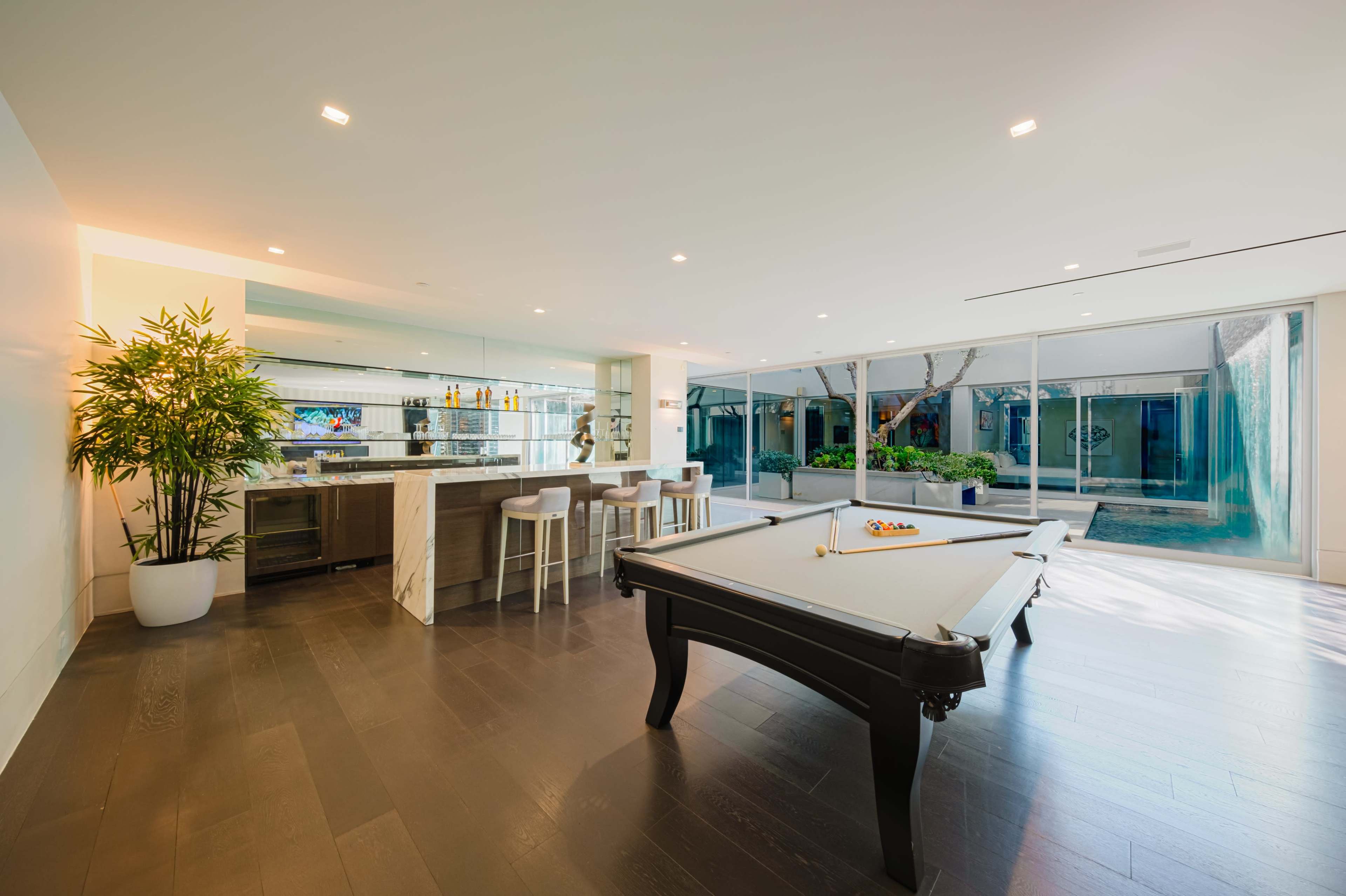 The image shows a modern game room with a billiard table, a bar area with stools, and large glass windows overlooking a small outdoor space with greenery.