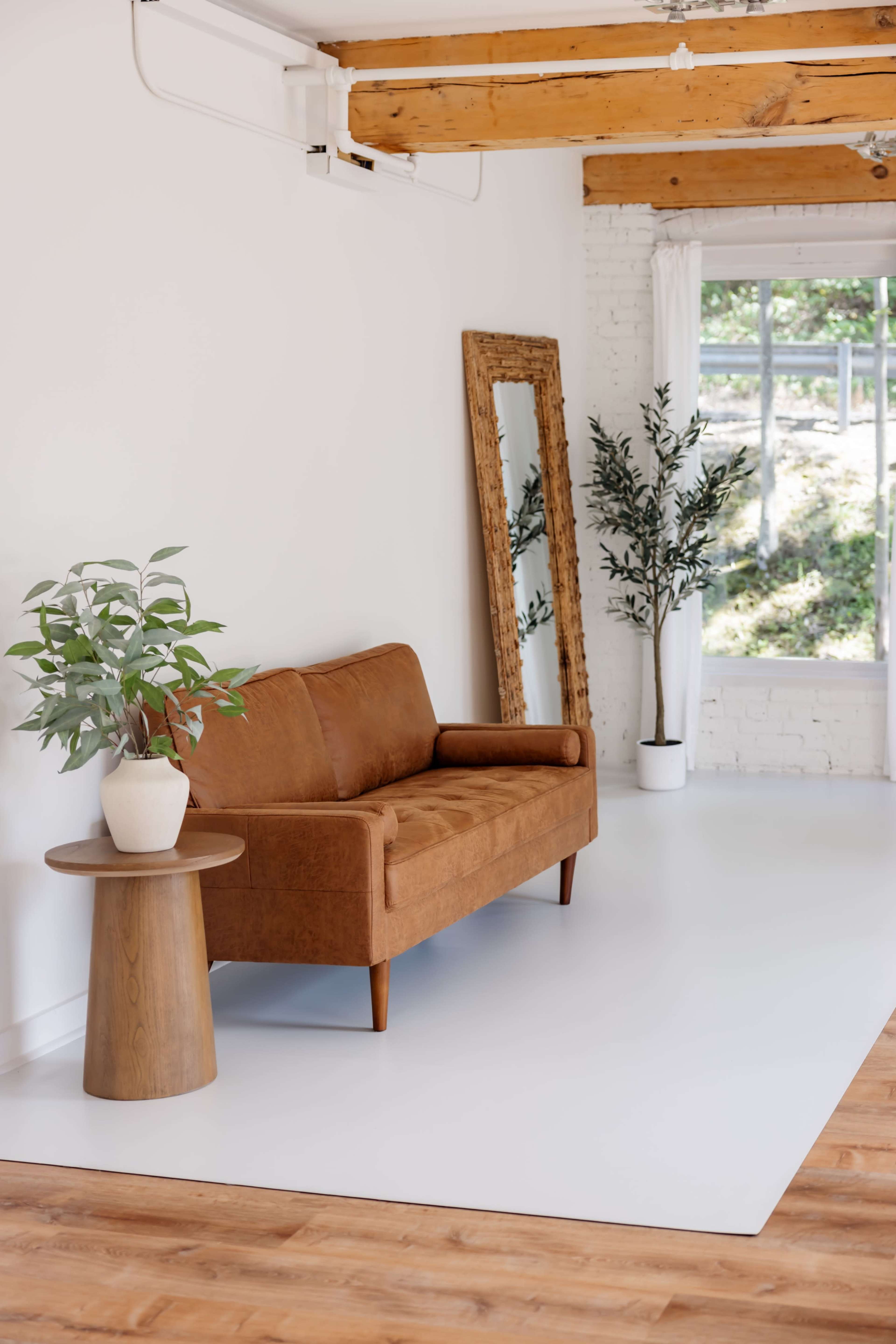 The image shows a minimalist interior space with a brown sofa, a round wooden side table, a potted plant, and a large mirror against a white wall, illuminated by natural light from a window.