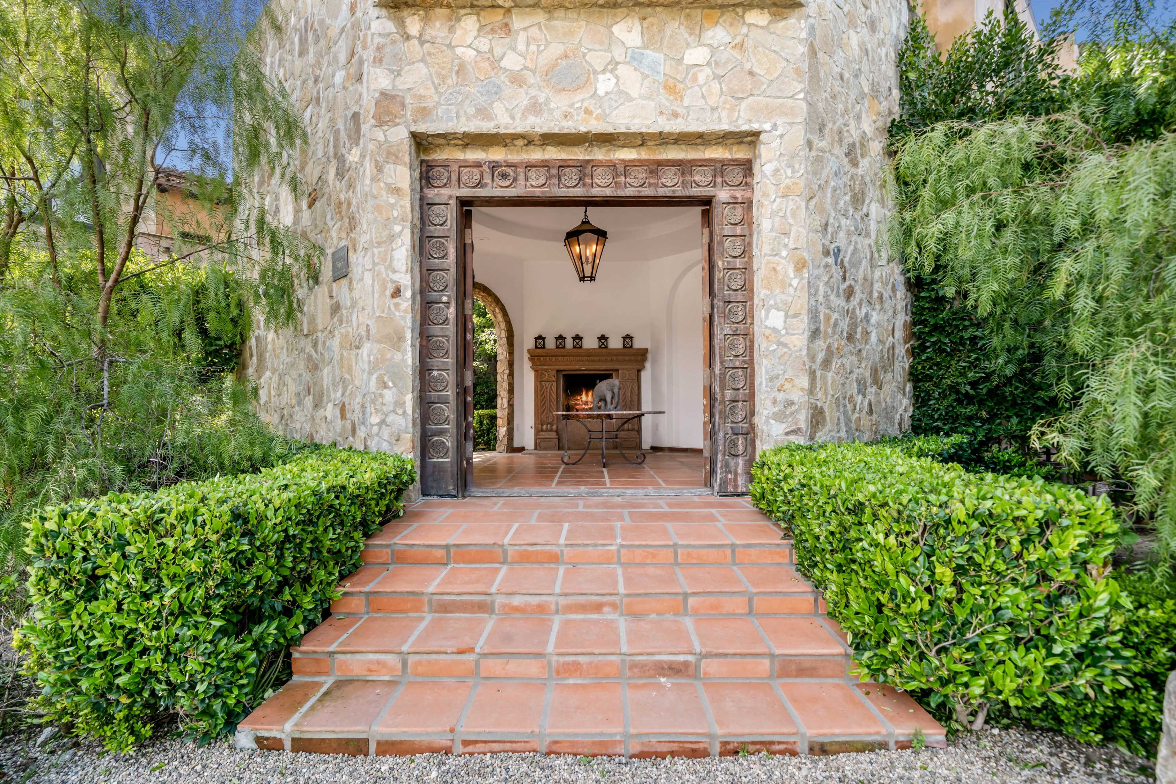 A stone entrance with a wooden door and a lantern leads to a room featuring a fireplace, flanked by neatly trimmed hedges and a tiled pathway.