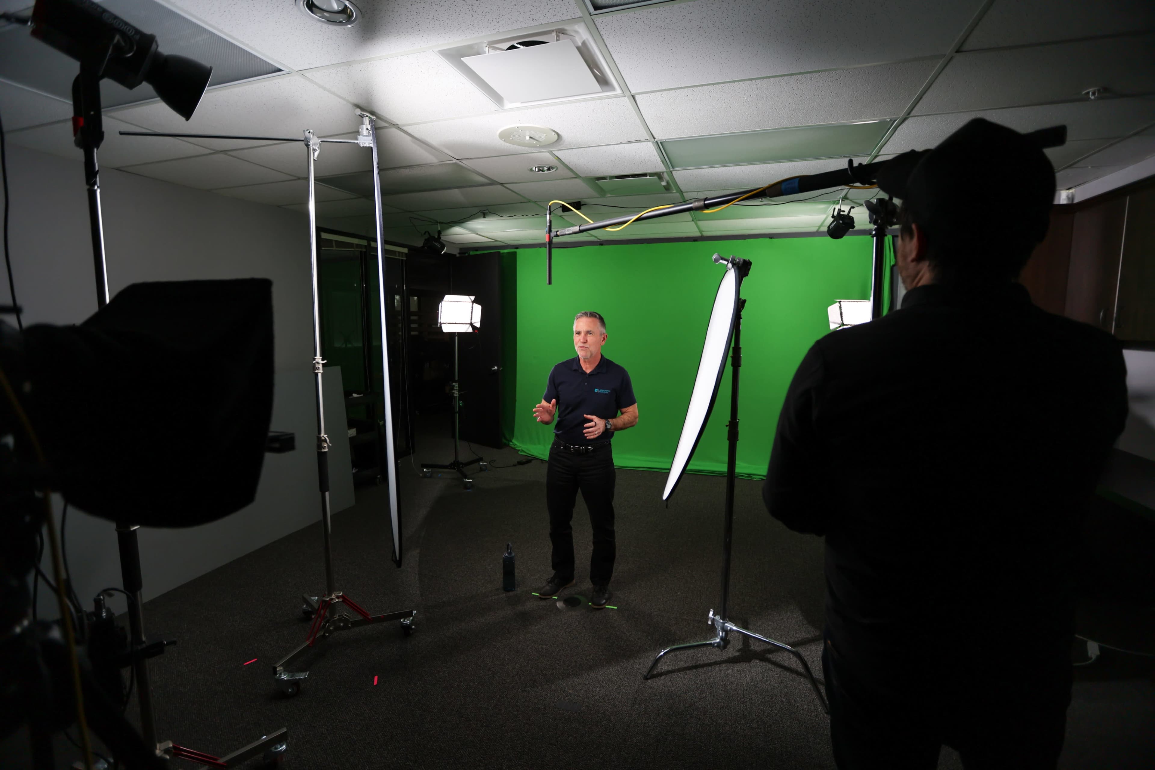 A man stands in front of a green screen while being filmed, surrounded by lighting equipment and a camera setup.