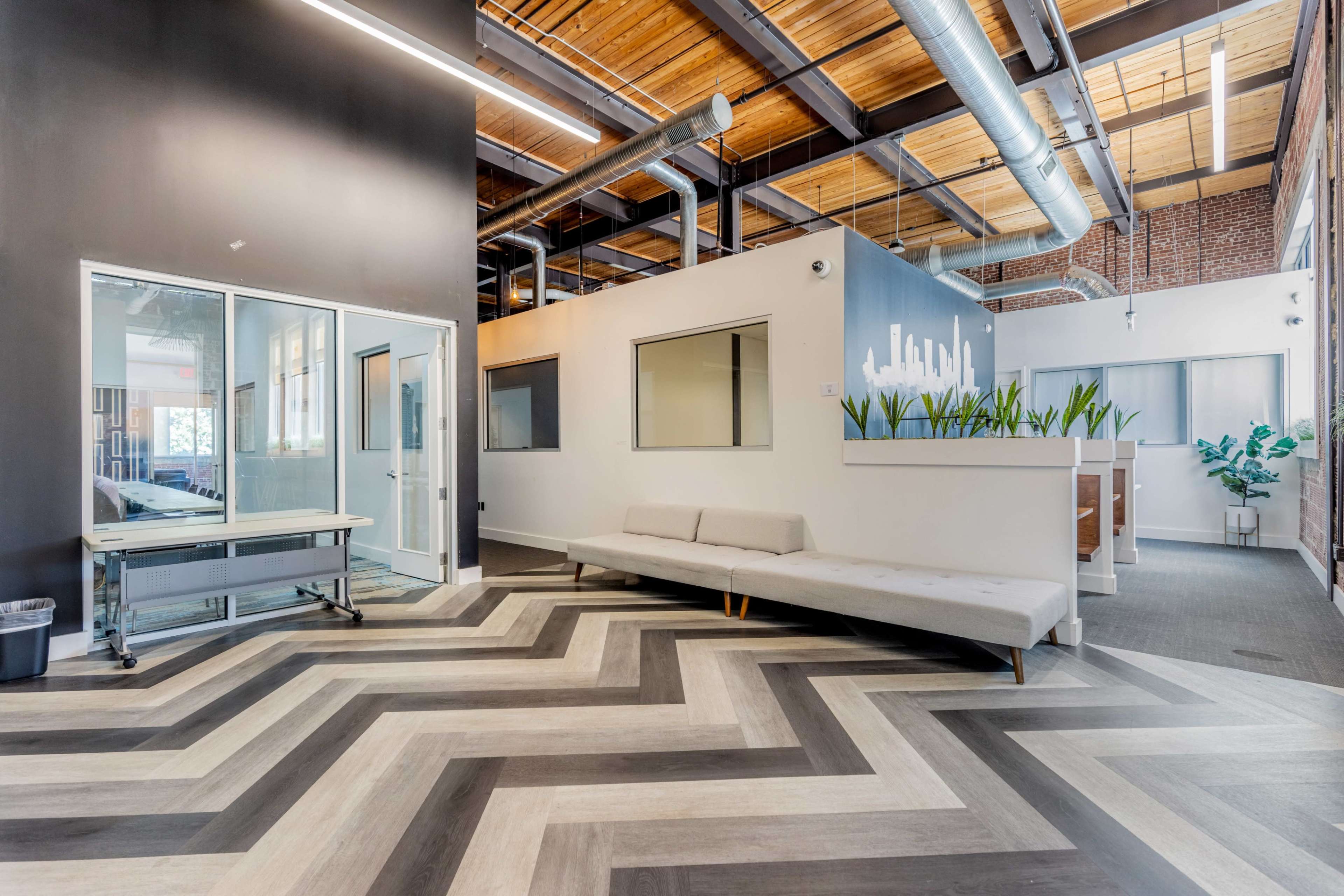 The image shows a modern office space featuring a chevron-patterned floor, a light gray sofa, and large windows along with greenery and various workstations.