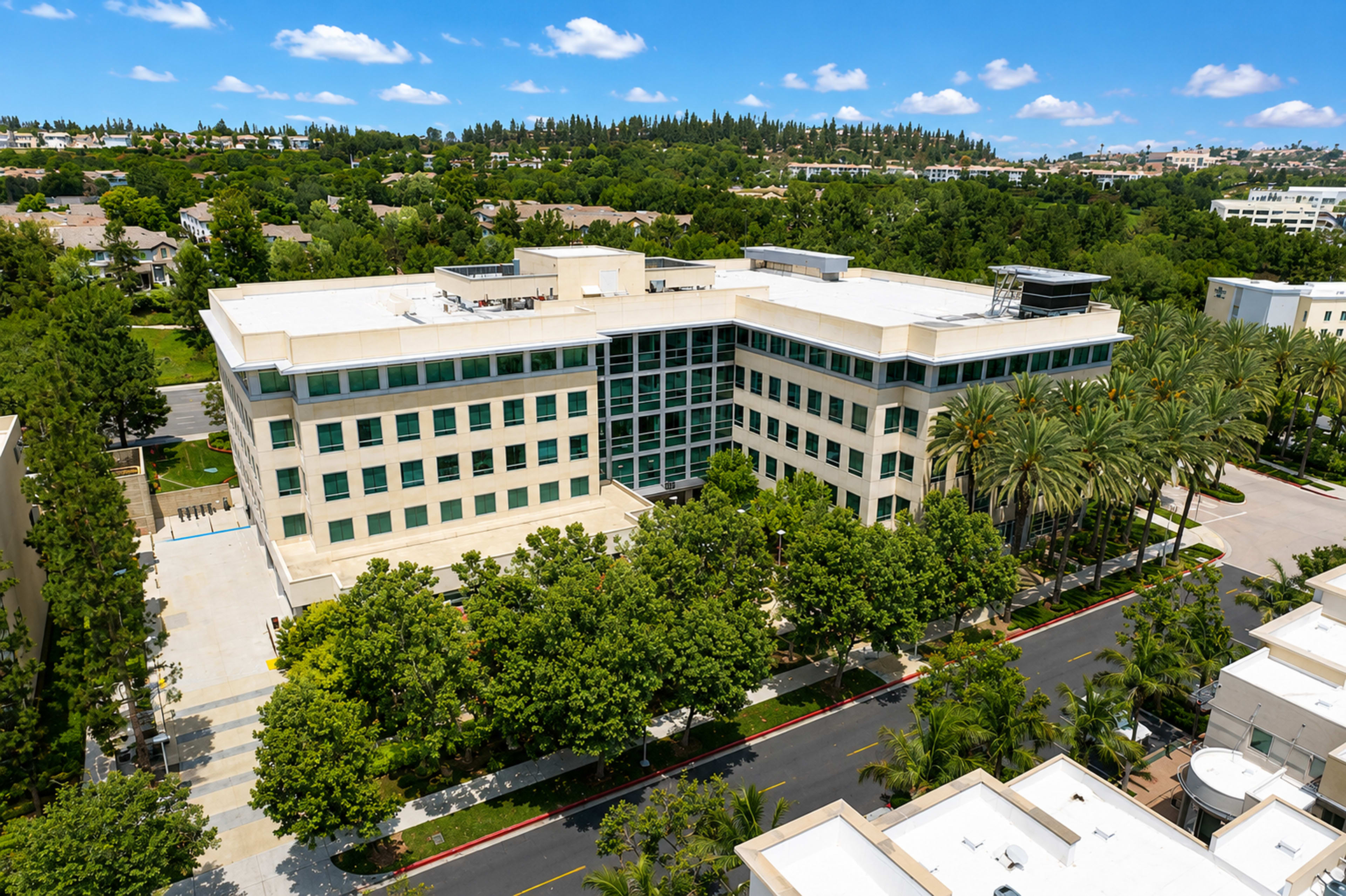 The image shows a modern multi-story building surrounded by neatly landscaped greenery and palm trees, with a paved road in front.