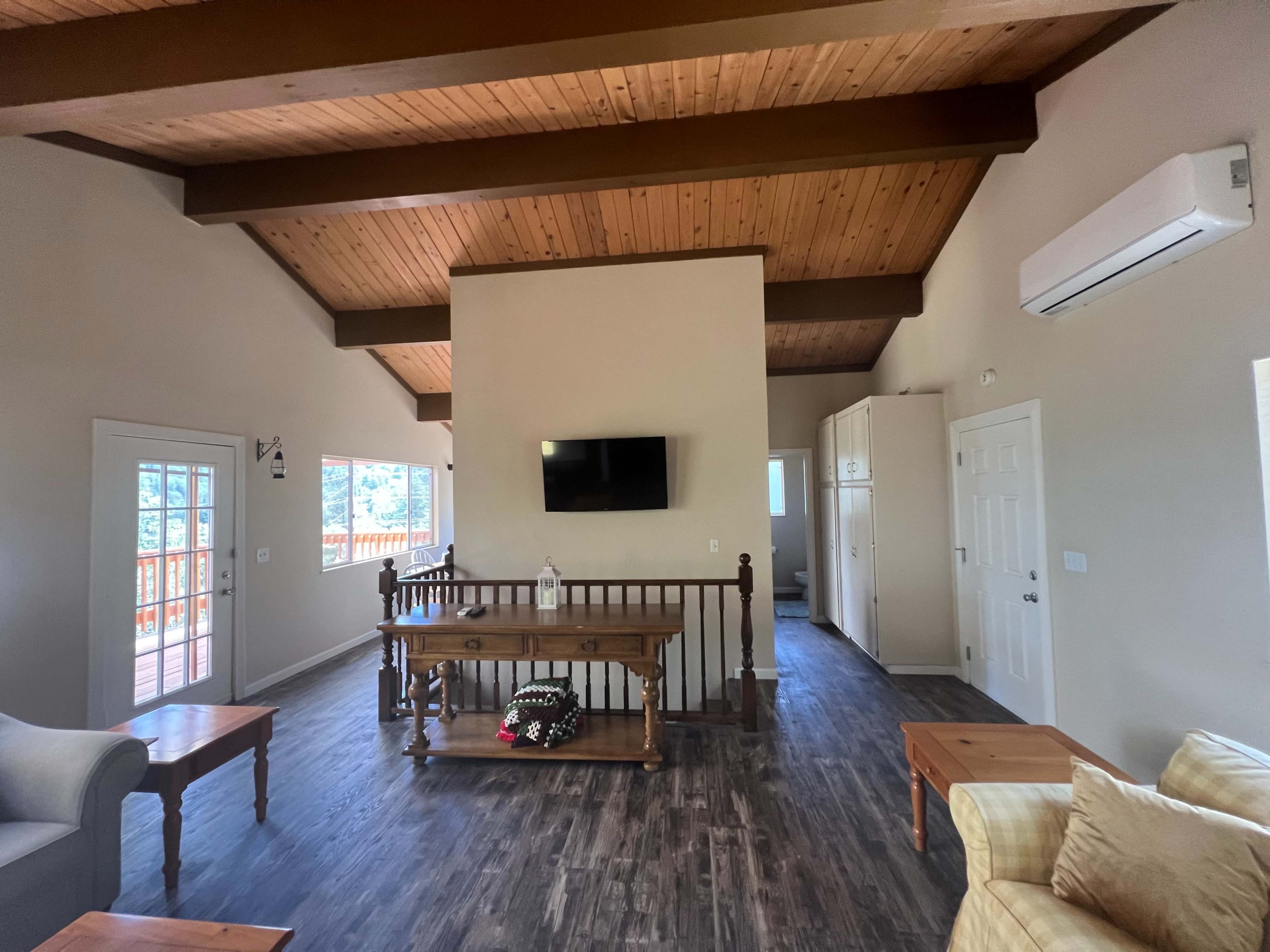 A living area with wooden ceilings, a flat-screen TV mounted on the wall, and adjacent rooms visible, including a kitchen area.
