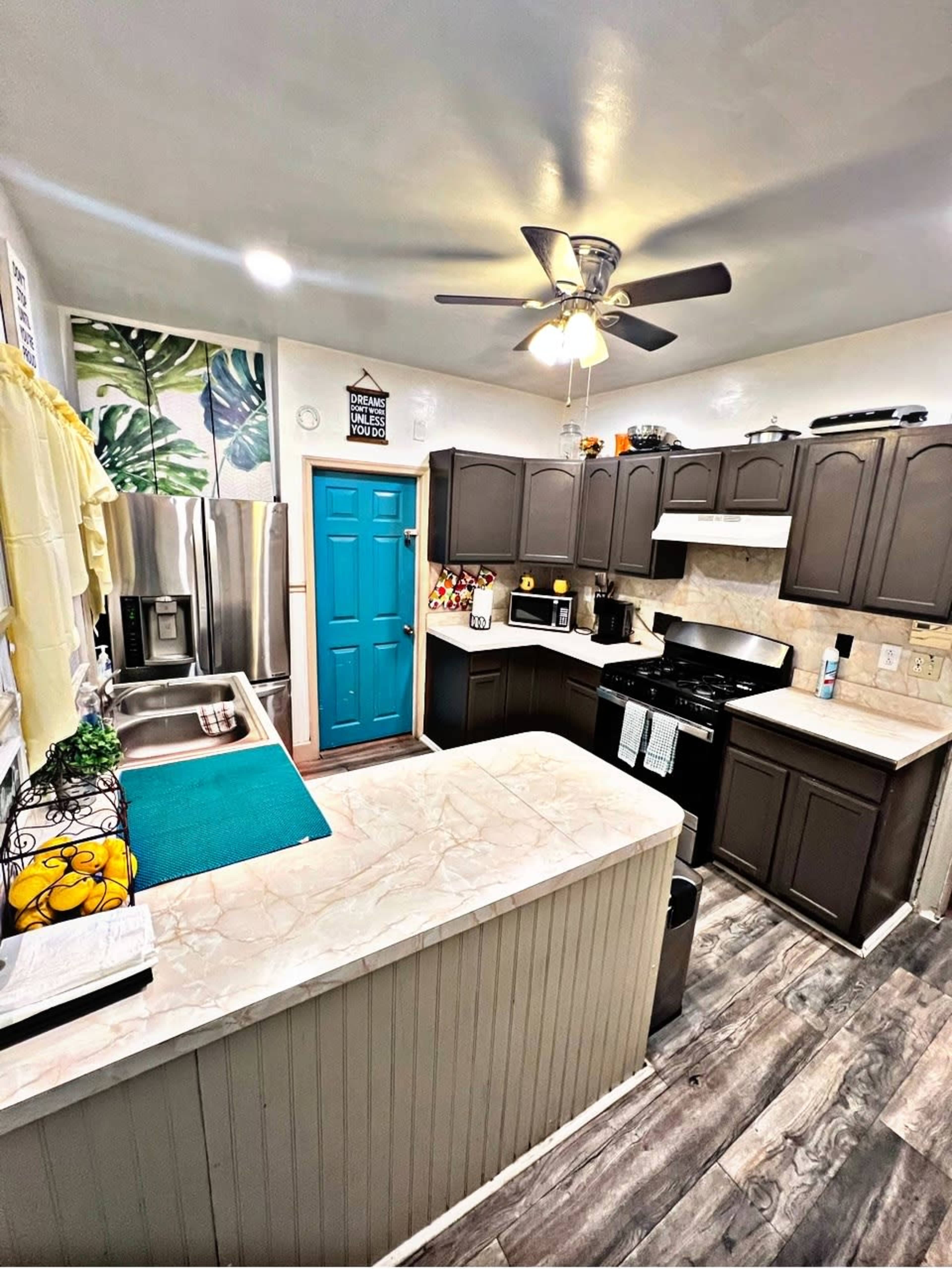 The image shows a kitchen with dark cabinets, a stainless steel refrigerator, and a blue door, featuring a marble countertop and a ceiling fan.