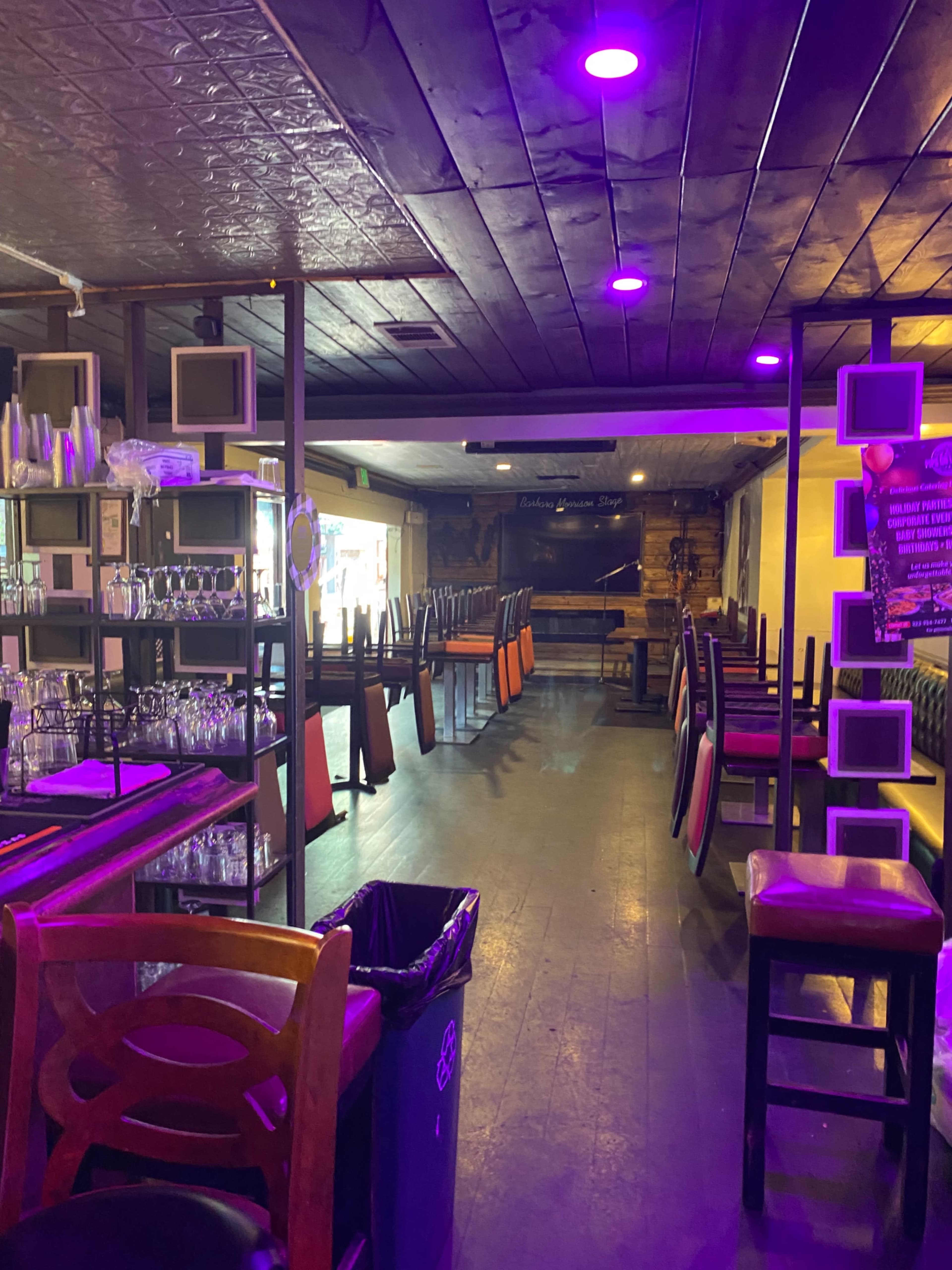 The image shows an empty bar area with tables and chairs stacked upside down, illuminated by purple lights.