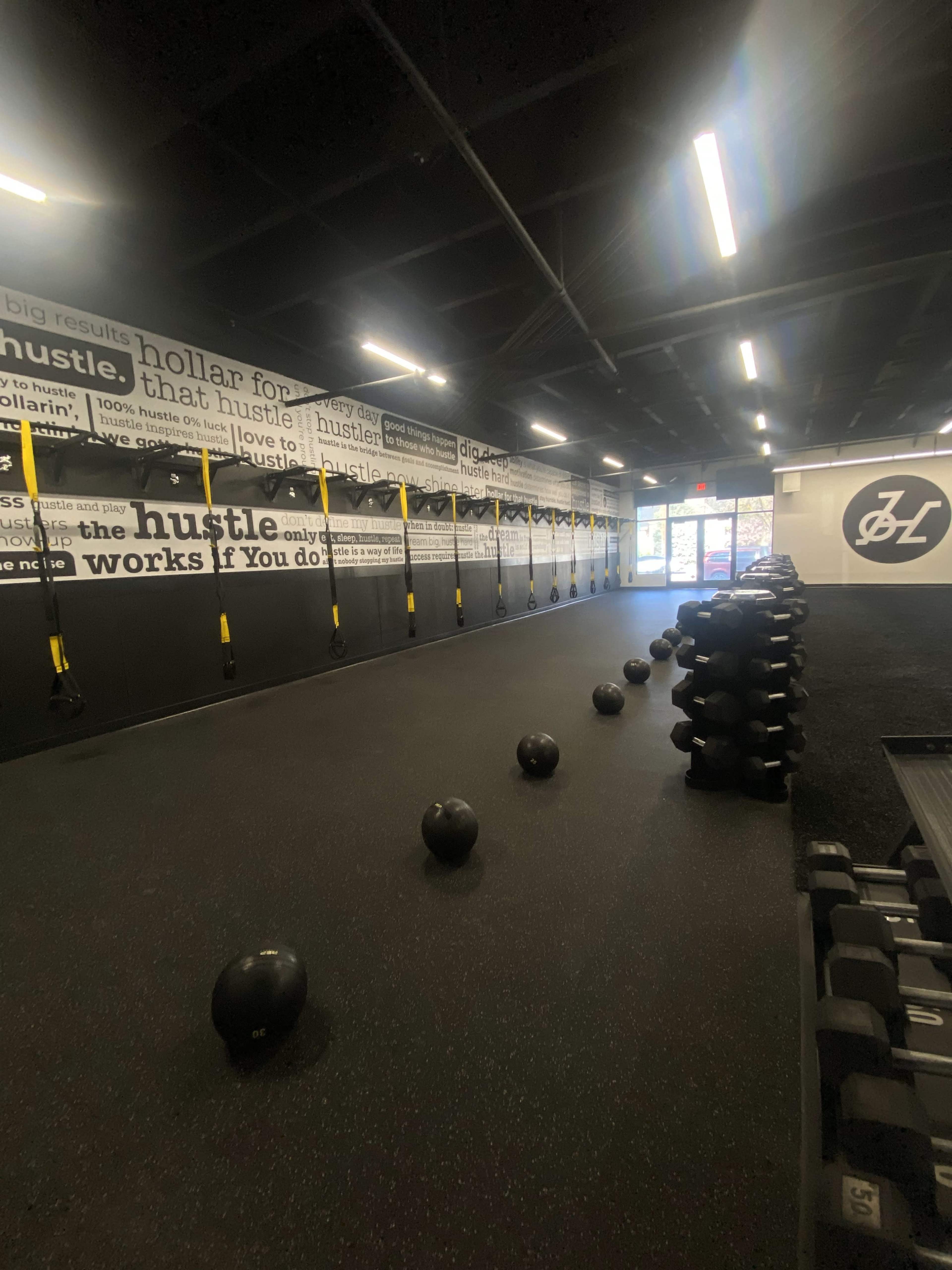 A fitness studio with medicine balls and dumbbells arranged on the floor, and wall-mounted resistance straps along the black walls decorated with motivational phrases.