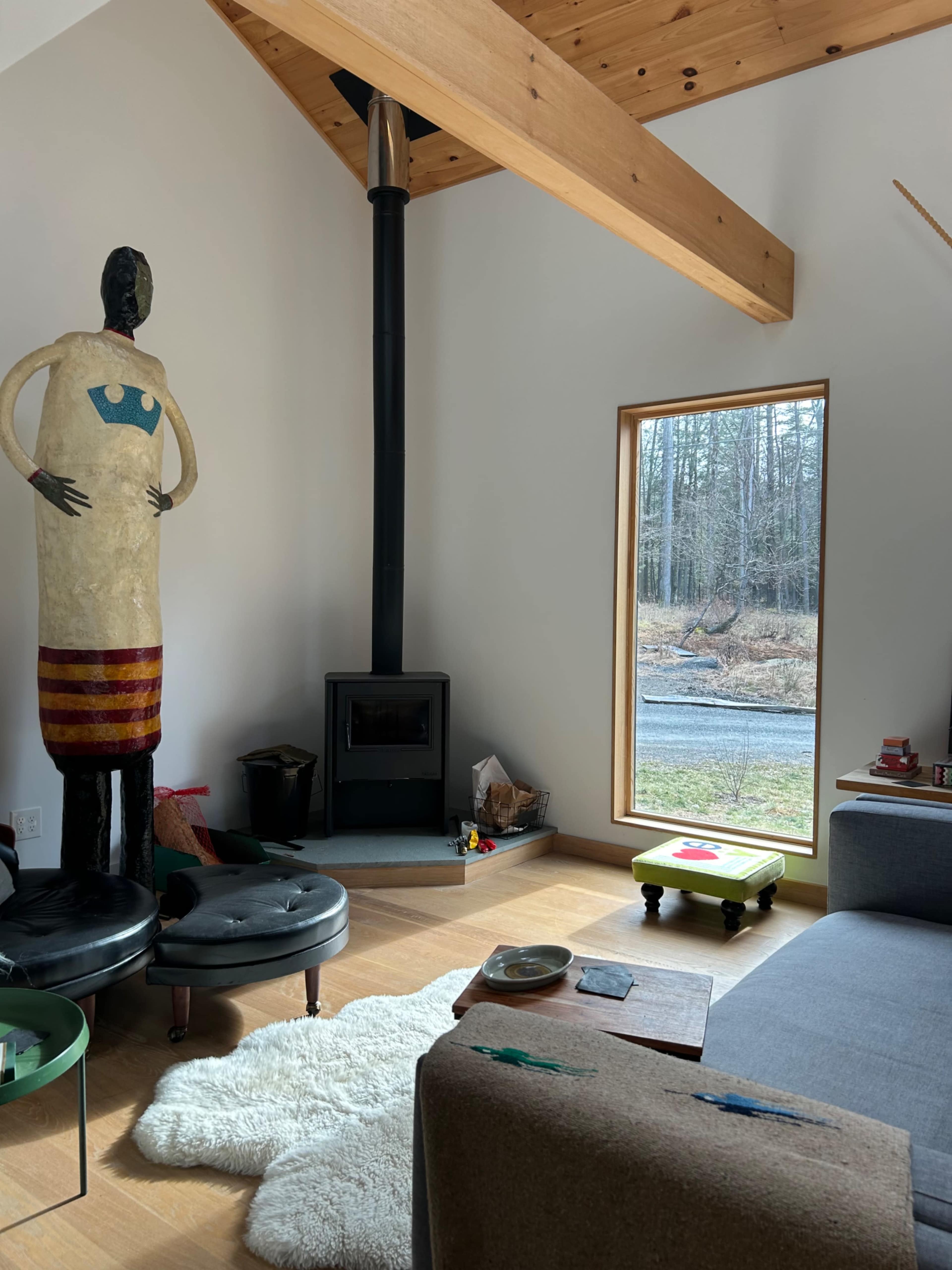 The interior of a modern living room features a tall, artistic figure next to a wood stove, with a large window showcasing an outdoor view.