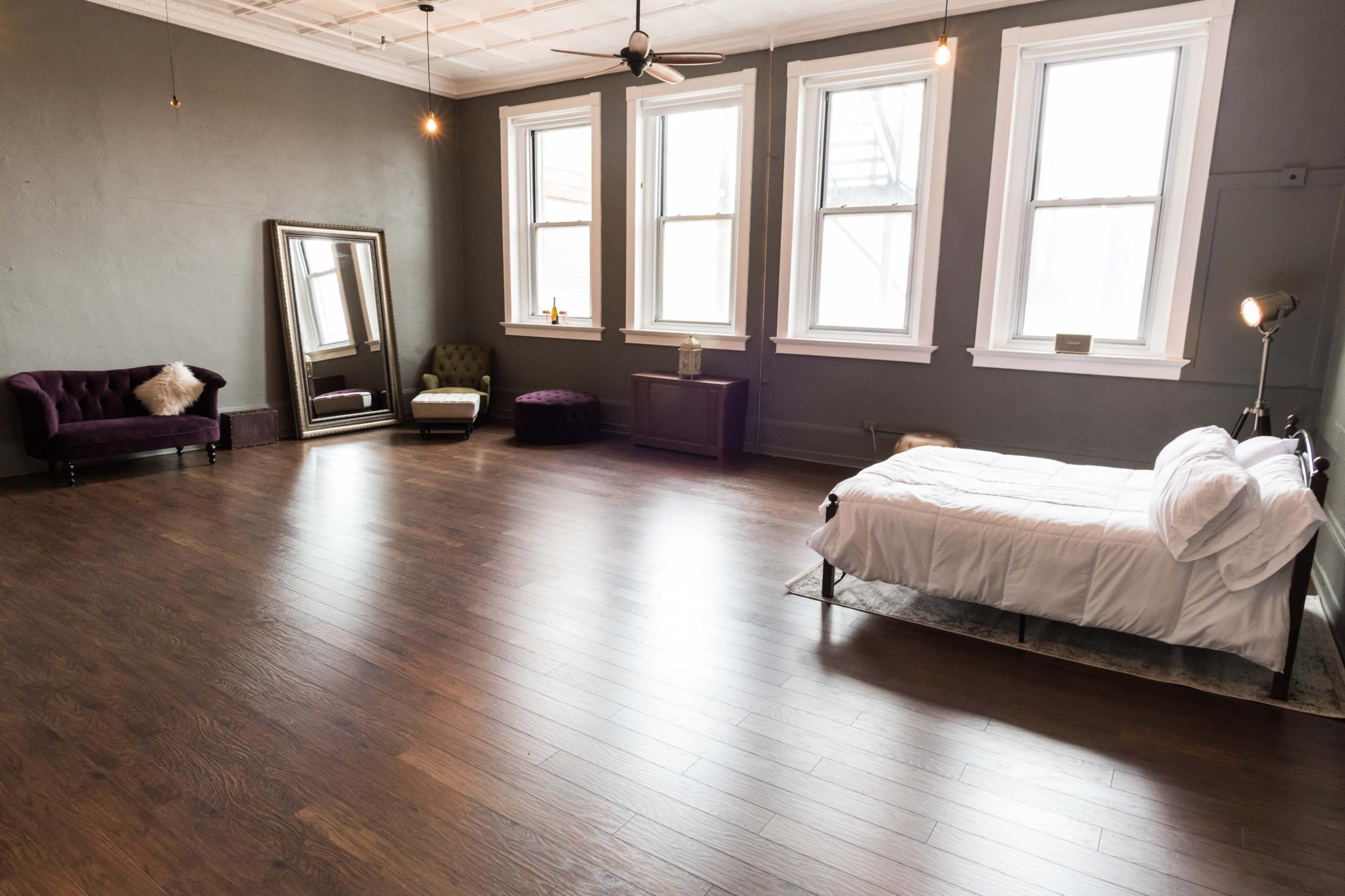 A spacious room with large windows, a bed, a mirror, and a couple of seating areas, all featuring hardwood flooring.