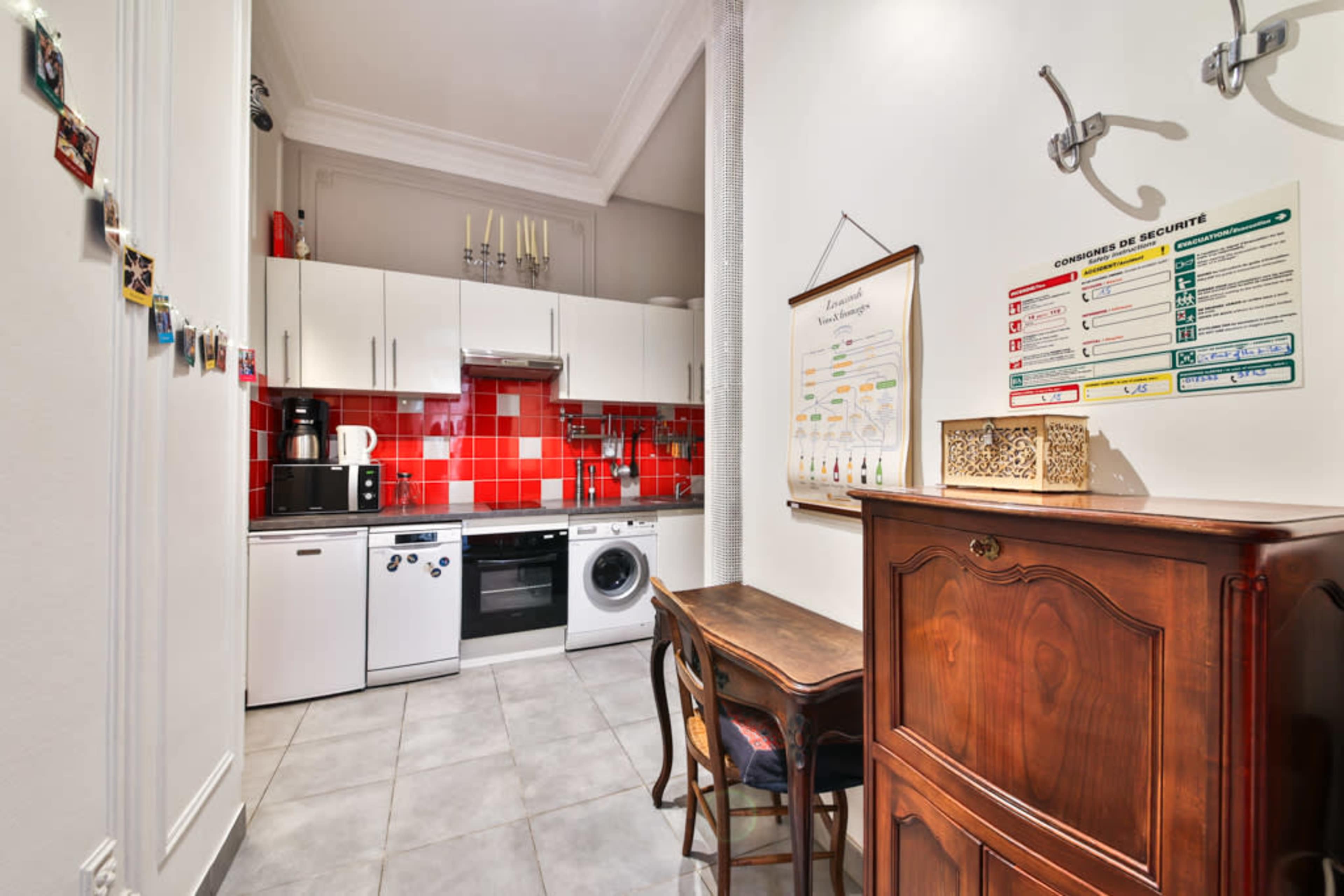 The image shows a small kitchen area with red tiled backsplash, white cabinetry, modern appliances, and a wooden table against the wall.
