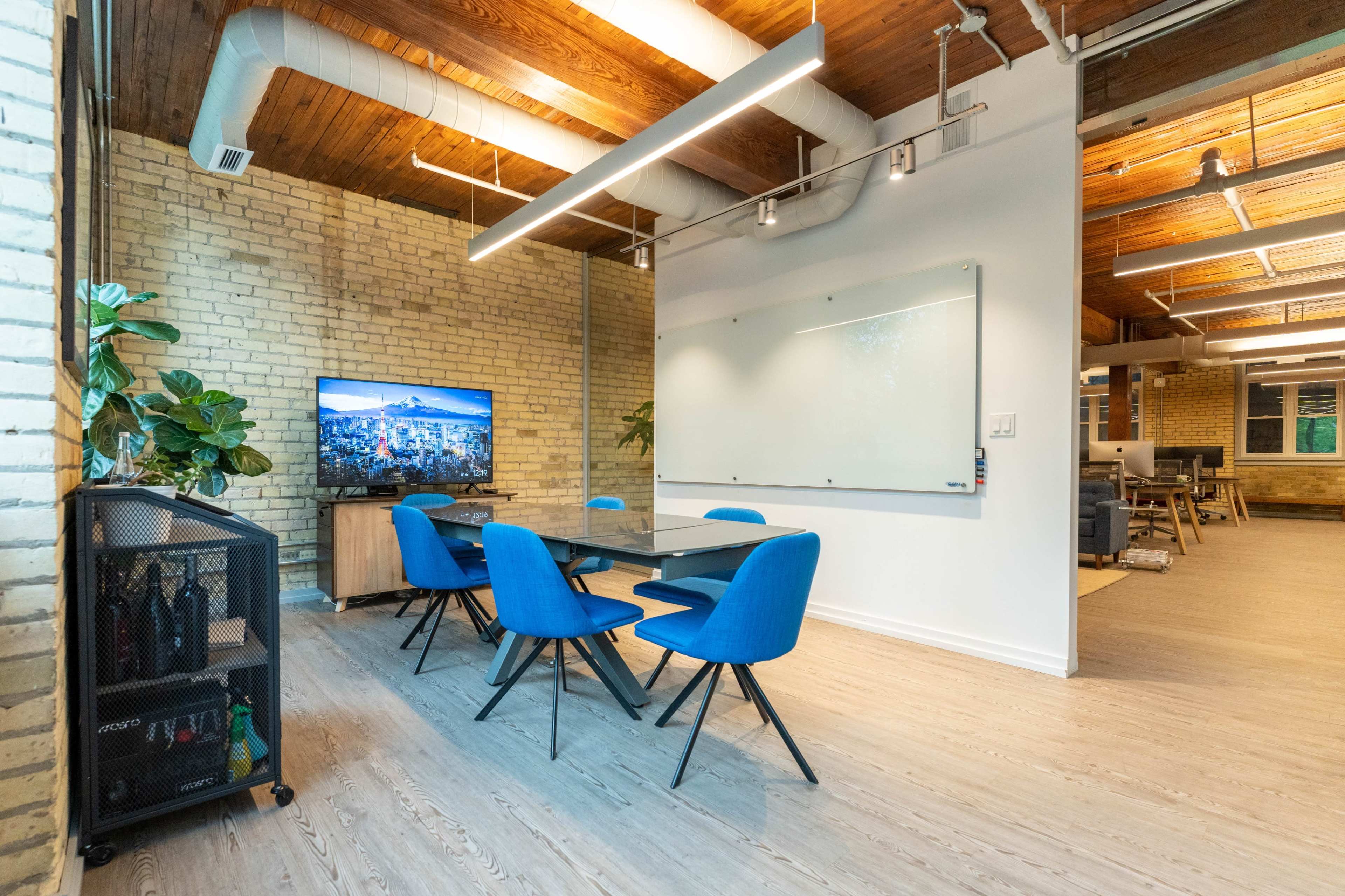A modern conference room features a large TV screen, a whiteboard, and a table surrounded by blue chairs against a backdrop of exposed brick and wooden beams.