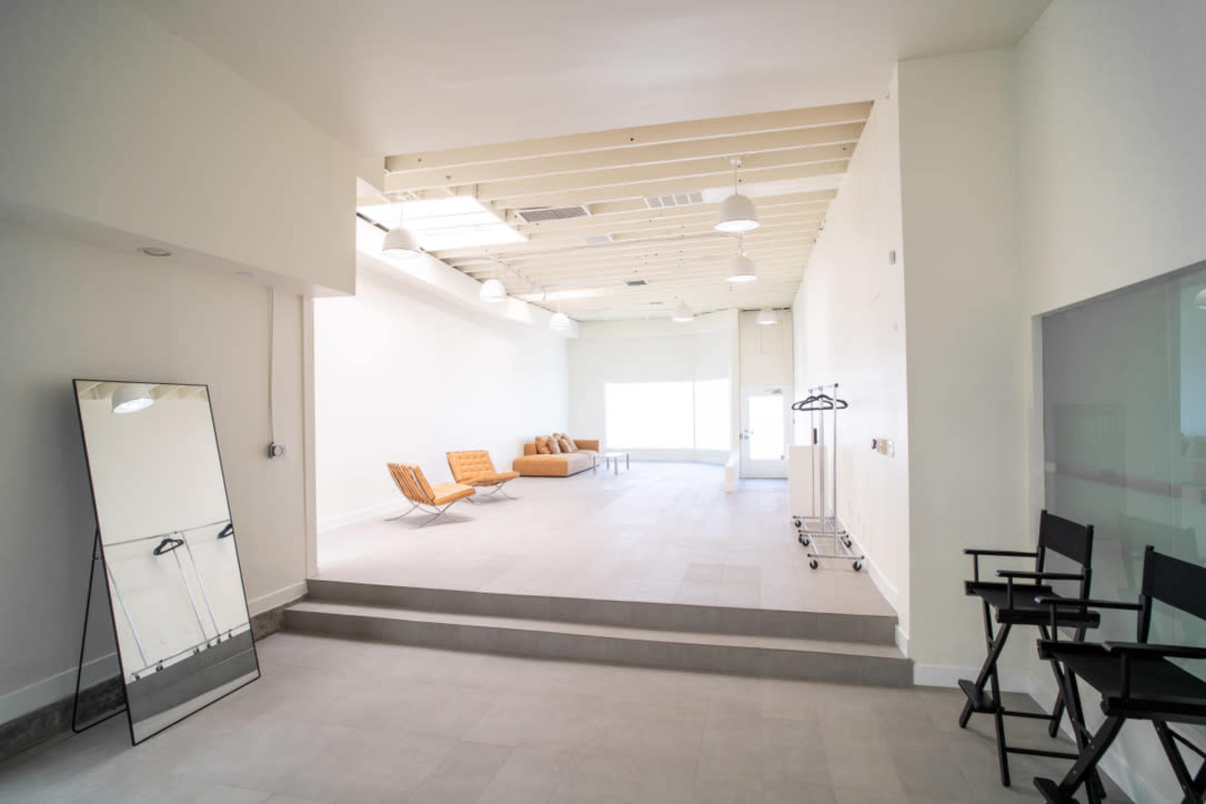 The image shows a bright, spacious interior with minimalist decor, featuring a large mirror, two orange chairs, and a few folding chairs along one side.