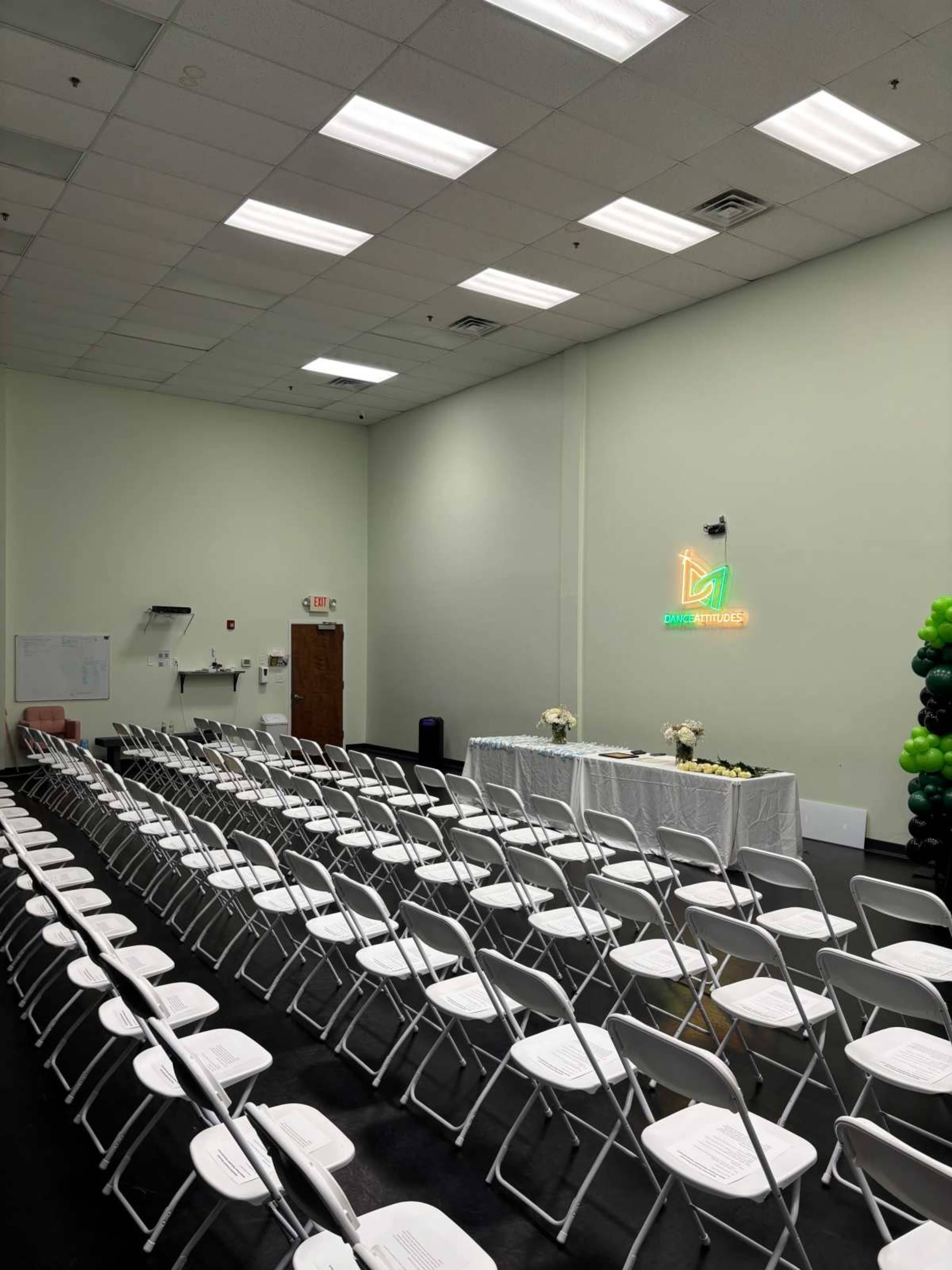 The image shows a neatly arranged row of white folding chairs facing a table decorated with flowers in a brightly lit room with balloons along one side.