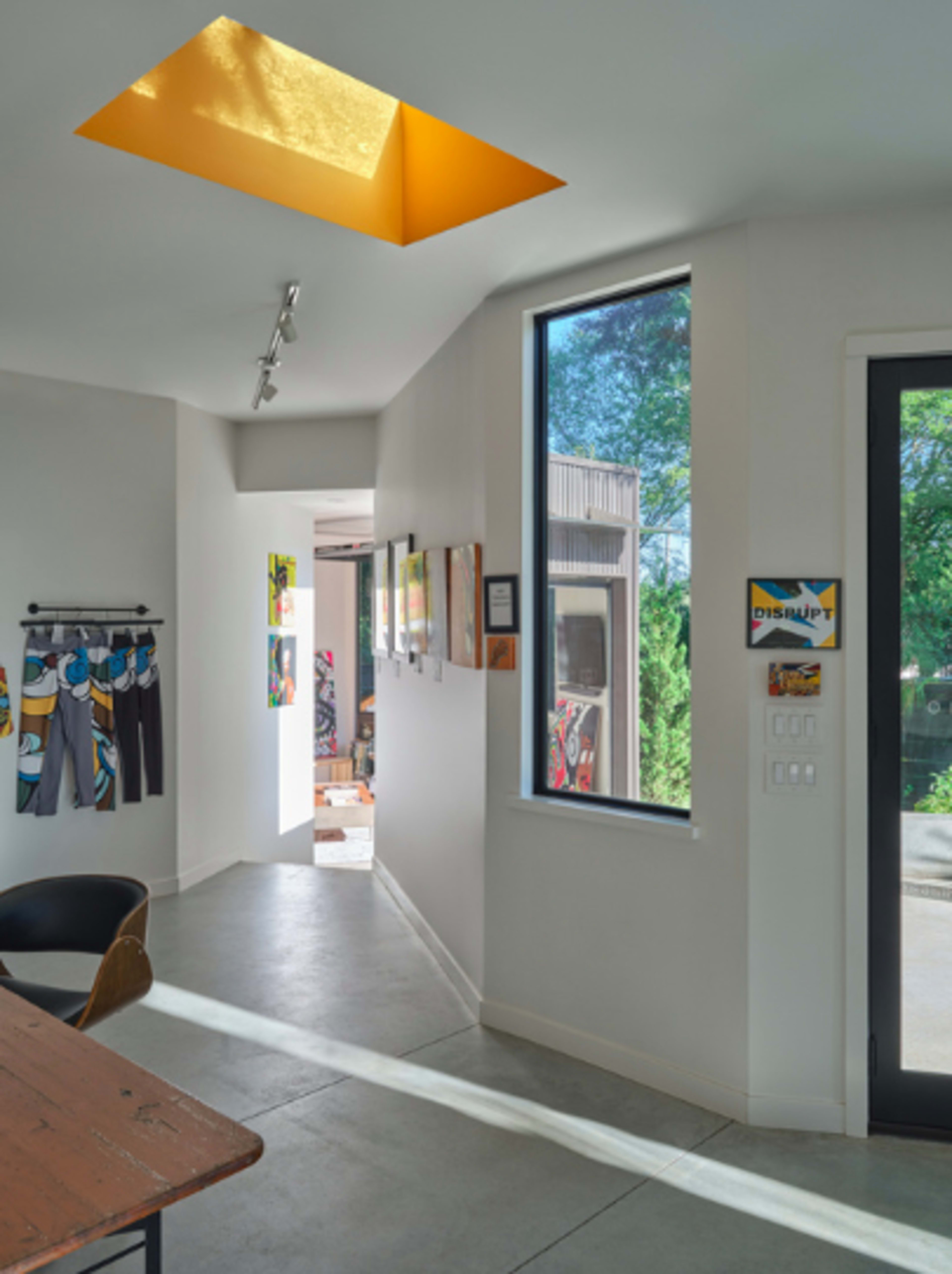 A hallway features a skylight and large windows, showcasing art on the walls and leading to another room.