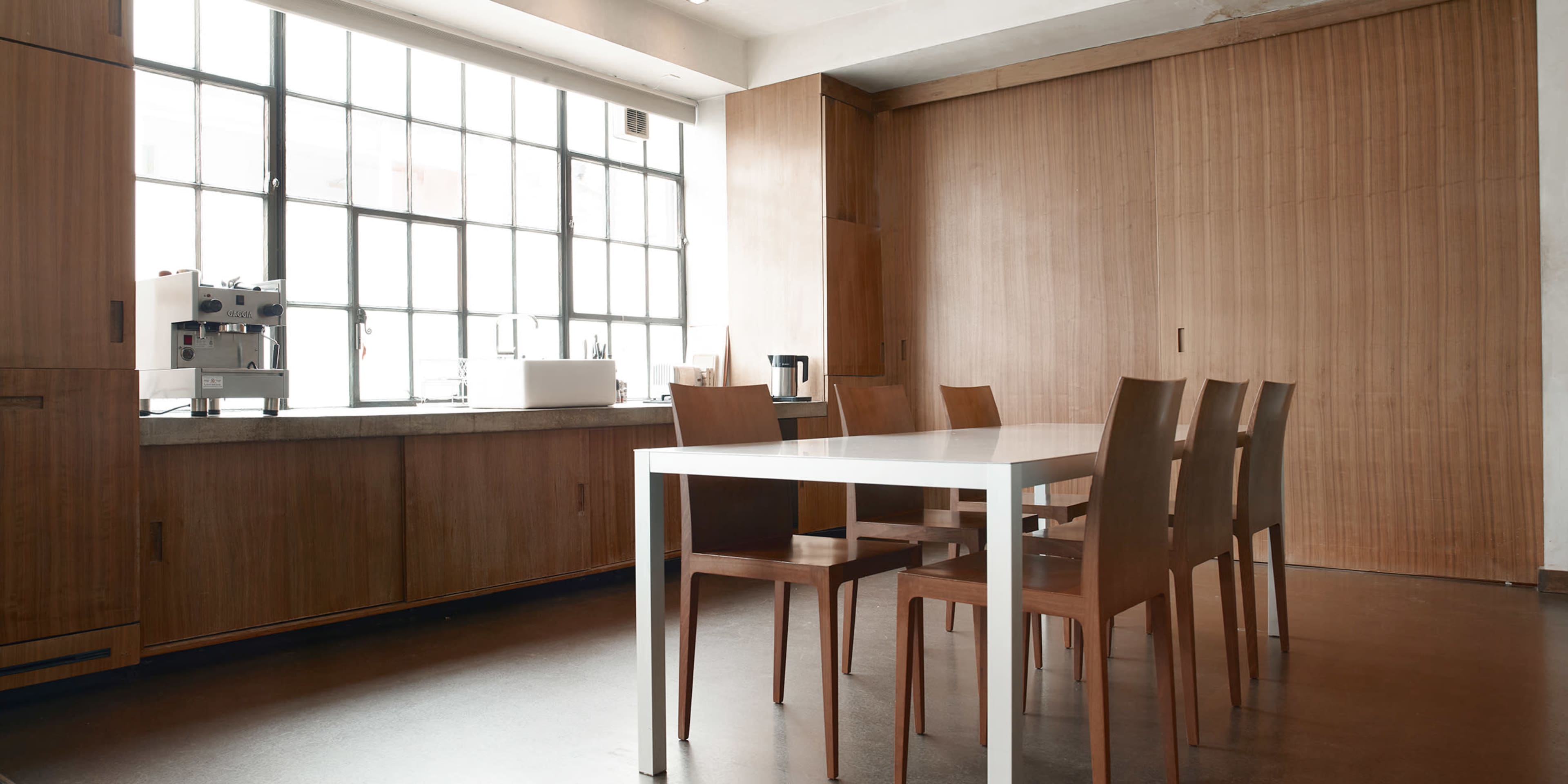 A modern kitchen features a large table surrounded by six wooden chairs and large windows that allow natural light to enter.