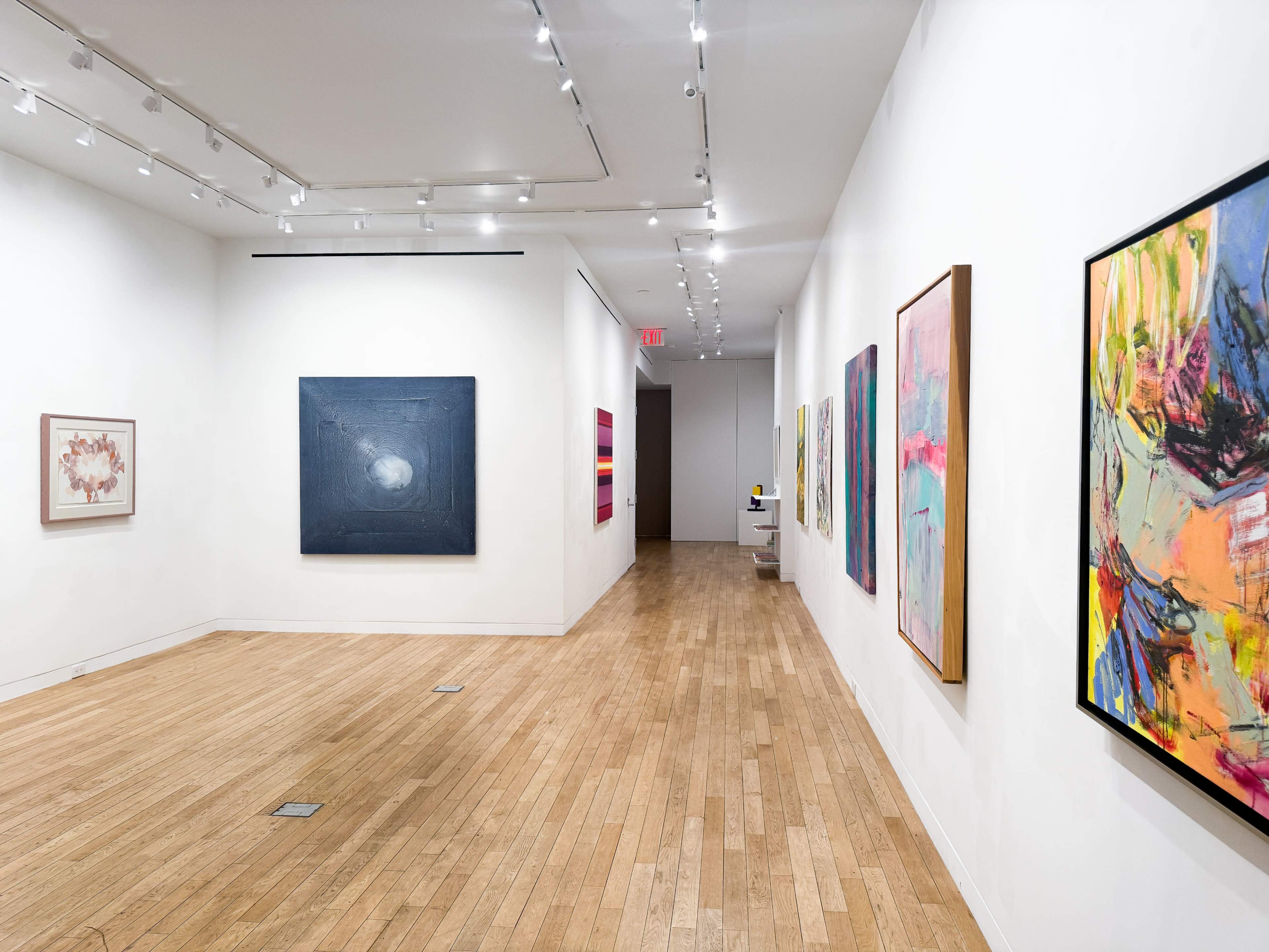A bright art gallery features a long hallway lined with various colorful abstract paintings on the white walls.