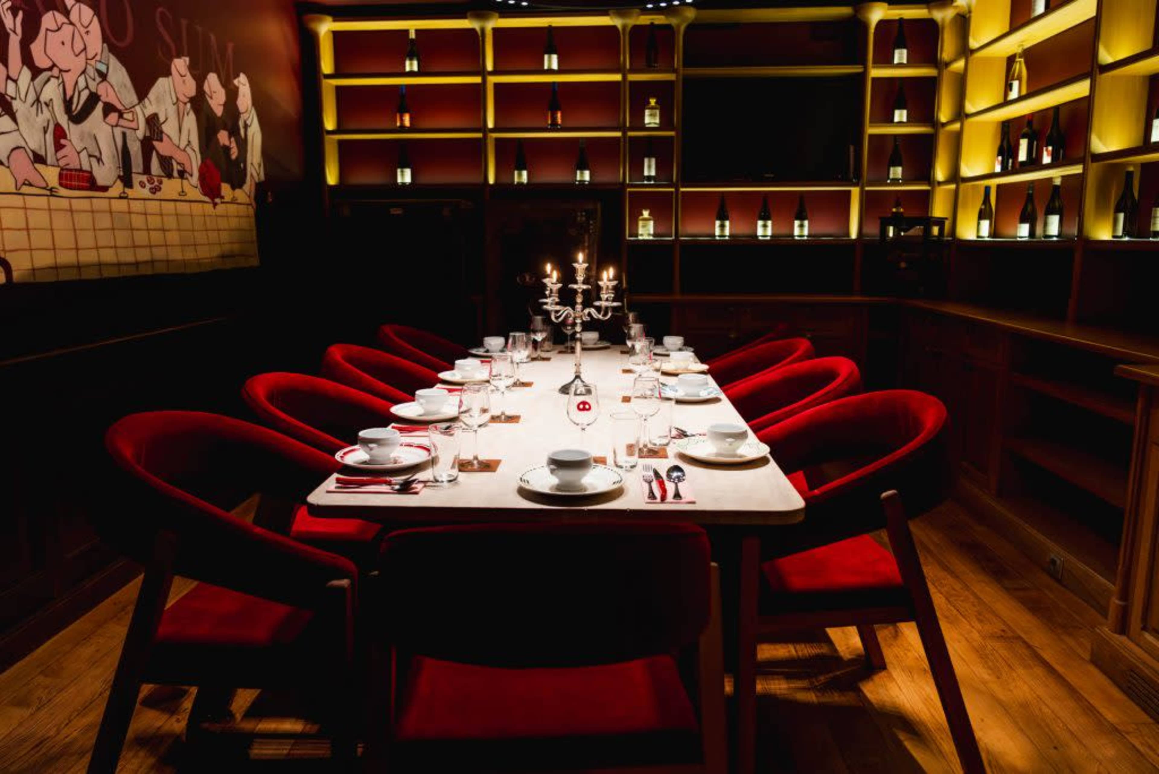 A long wooden dining table set with dishes and cutlery is surrounded by red upholstered chairs in a dimly lit restaurant.