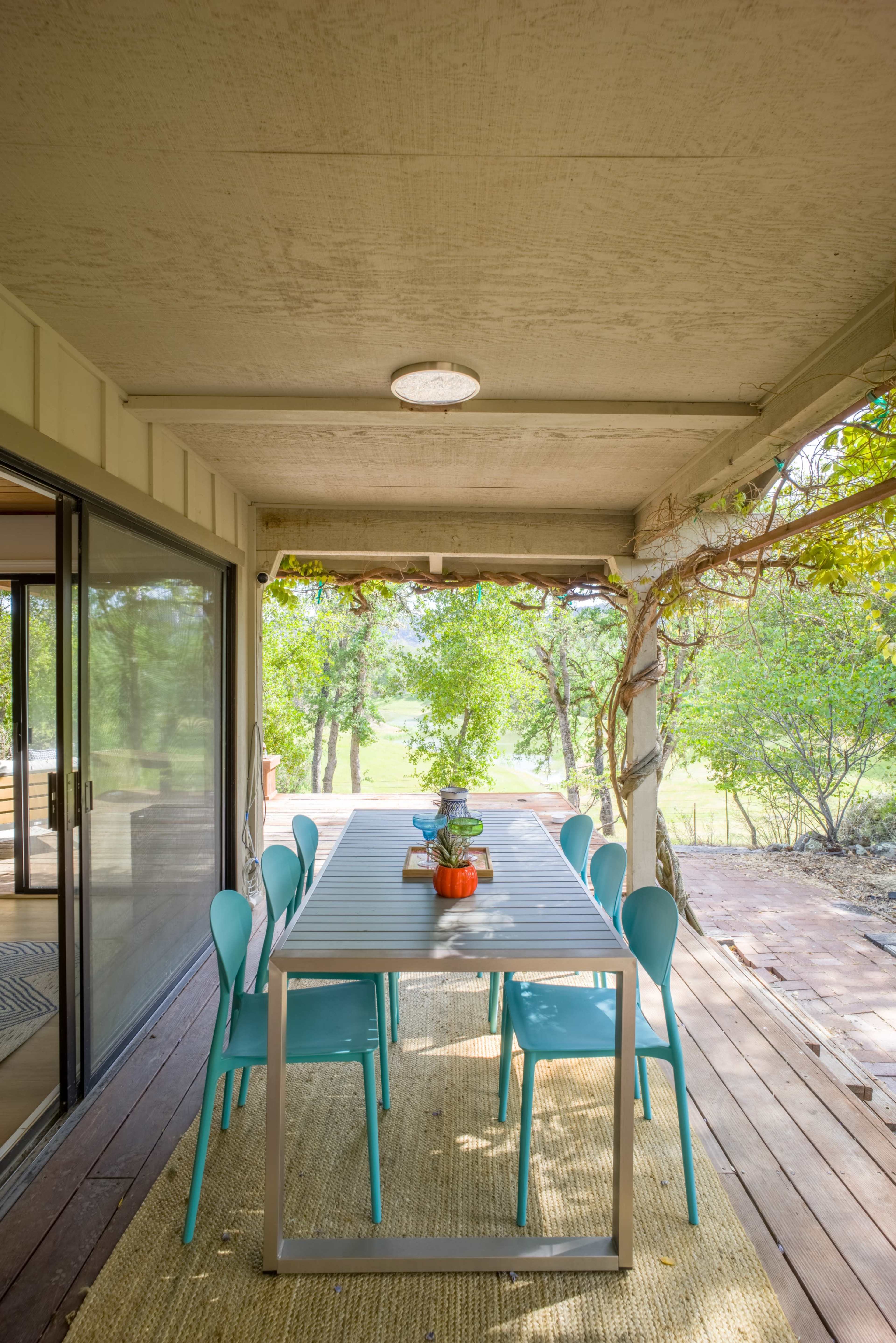 A long dining table with blue chairs is situated on a porch surrounded by greenery.