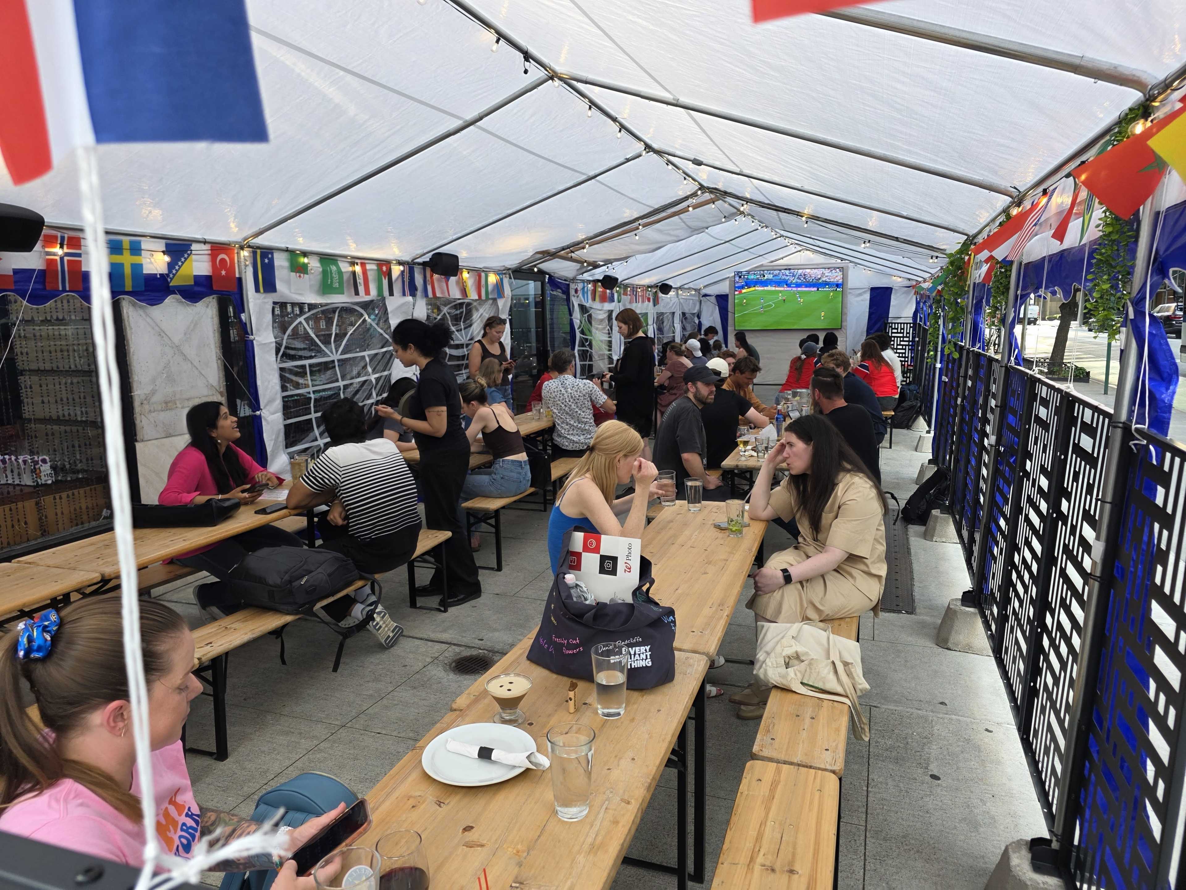 A gathering of people sits at long tables under a tent, watching a sports event on a large screen while flags from various countries hang overhead.