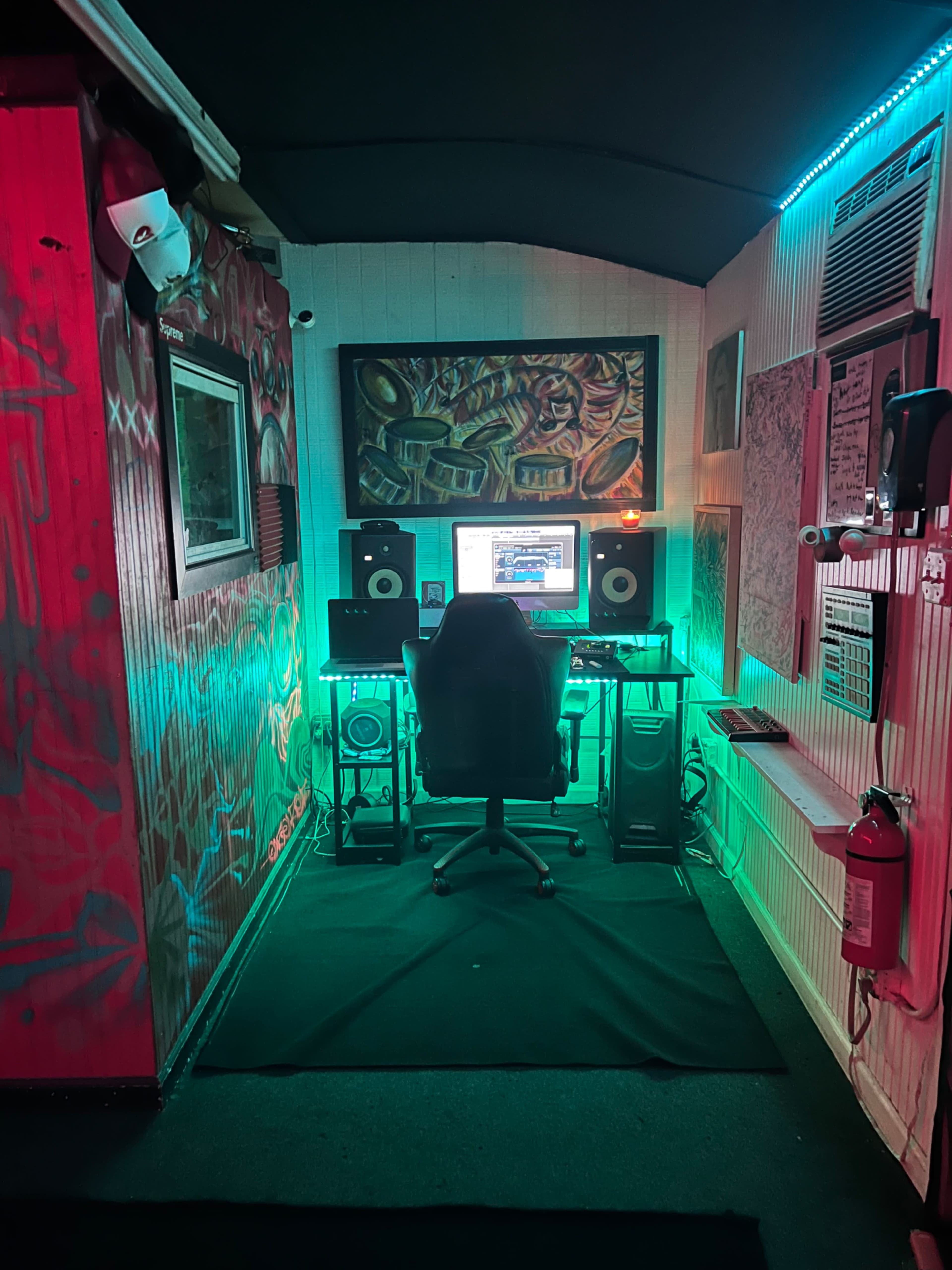 A small music studio features a central desk with an illuminated computer monitor, speakers, and a black office chair, surrounded by colorful graffiti on the walls.