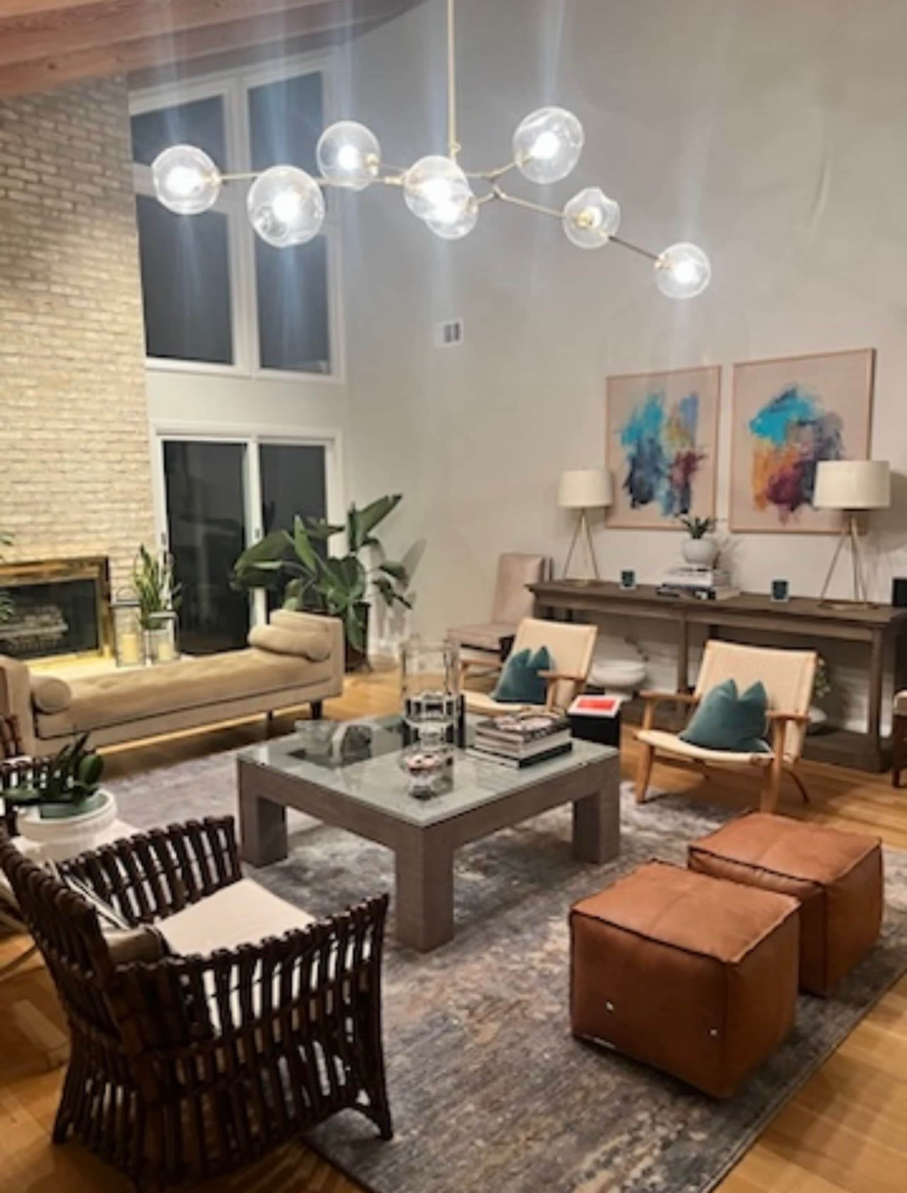 The living room features a light-colored sofa and armchairs arranged around a coffee table, with large windows and modern decor, including a stylish chandelier and artwork on the walls.