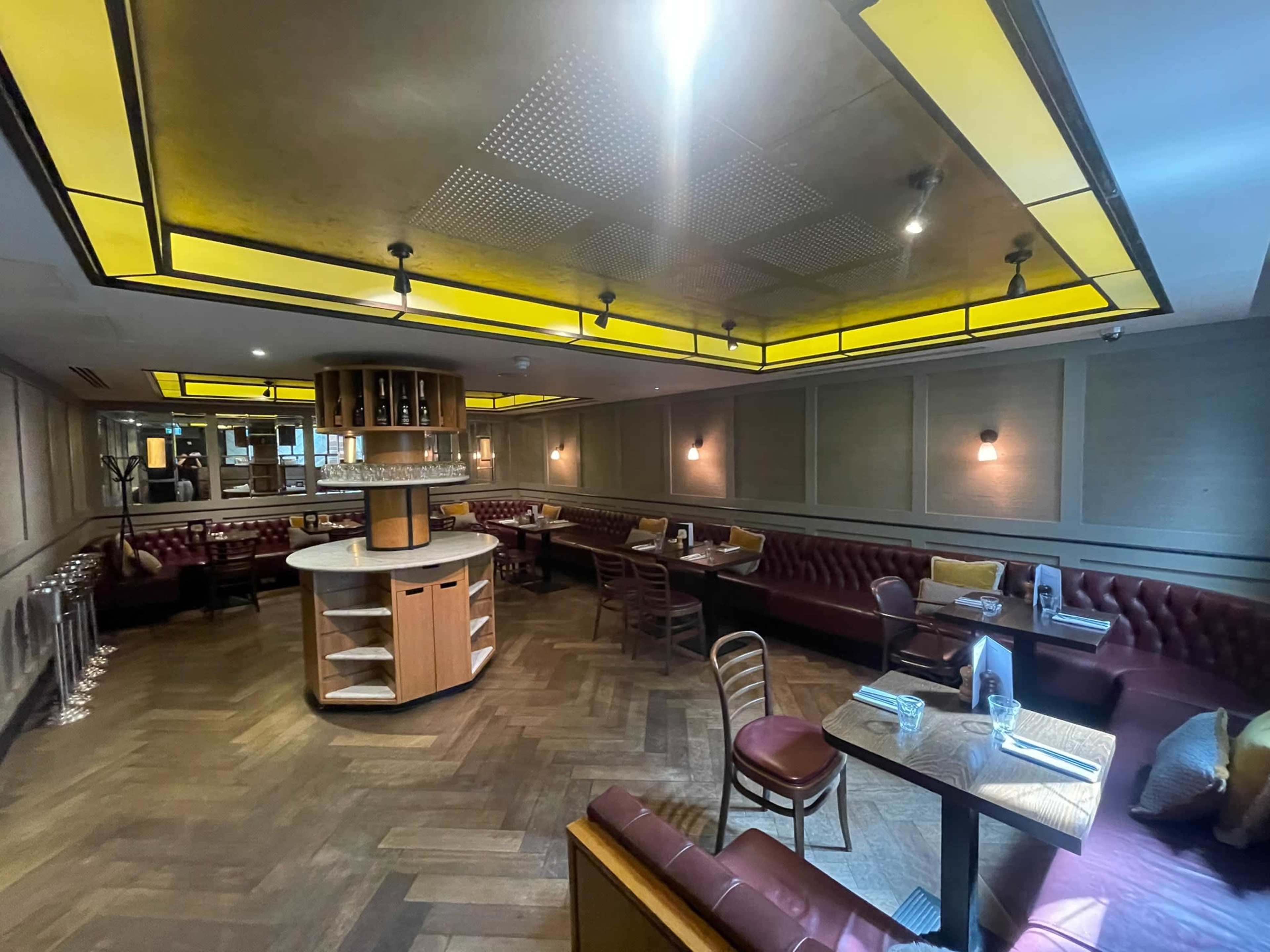 The image shows a modern restaurant interior featuring wooden flooring, burgundy leather seating, and a central circular dining area with tables.