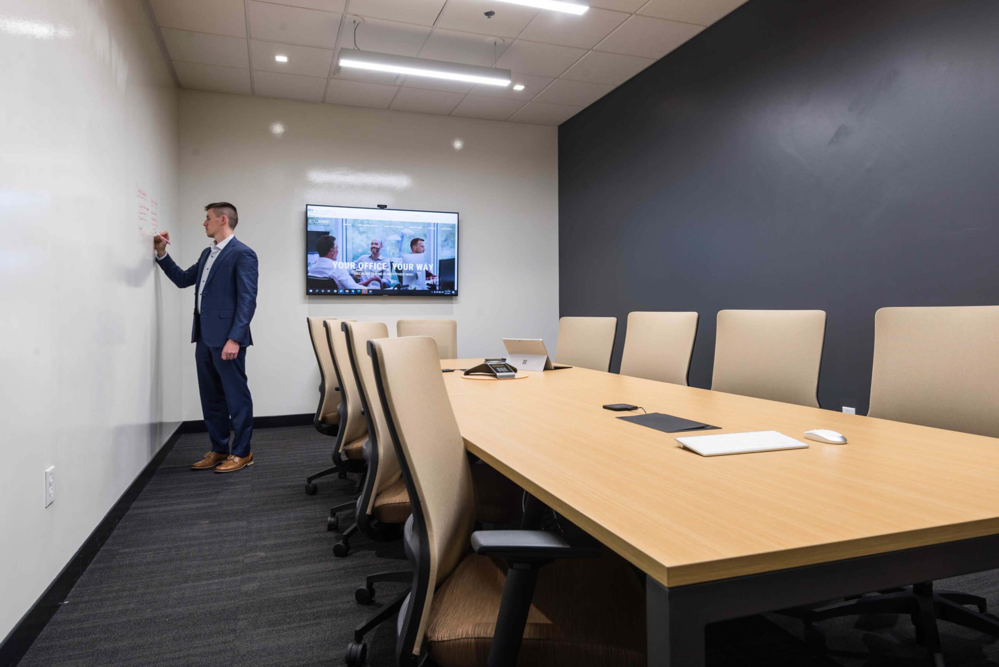 A man in formal attire writes on a whiteboard in a modern conference room equipped with a large table and a television screen.