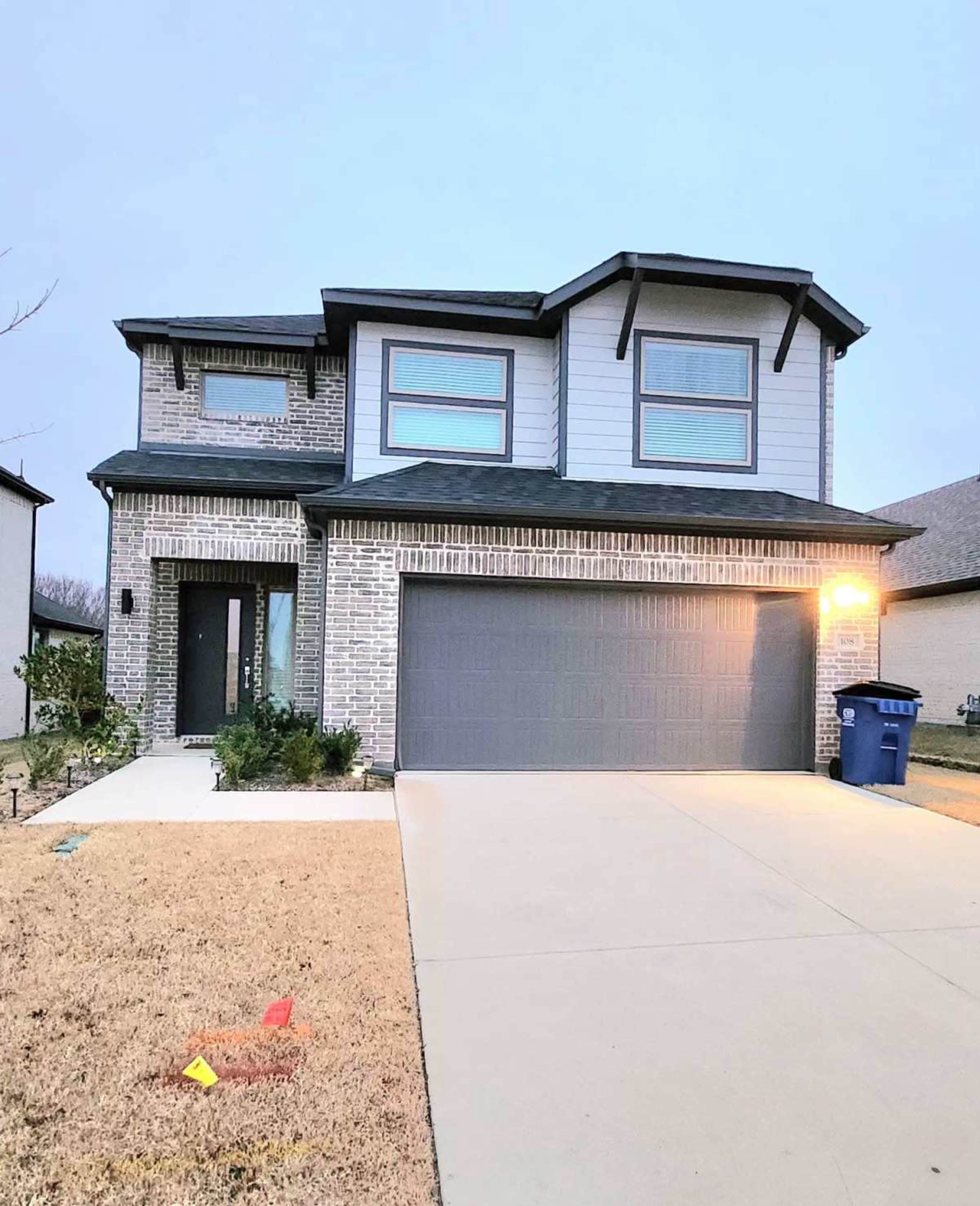 The image shows a two-story brick house with a gray garage door and a small front yard.