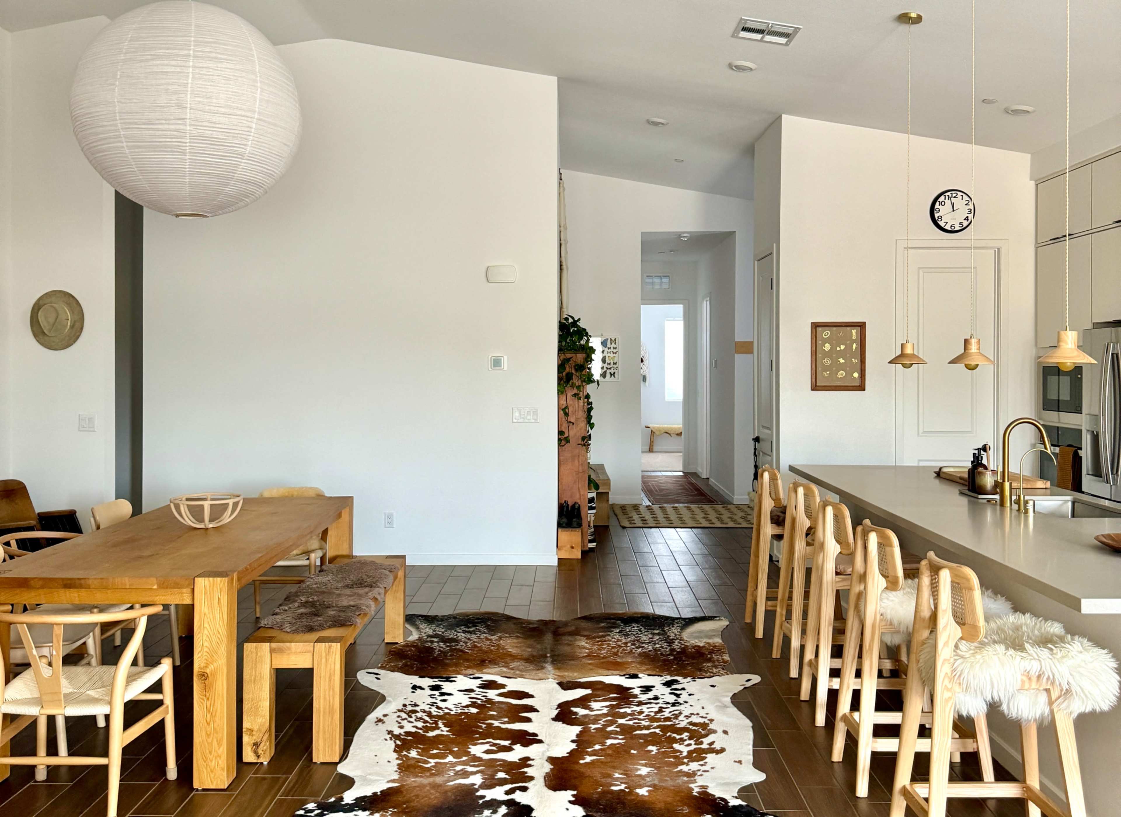 The image shows a modern dining area featuring a wooden table with chairs, a kitchen island with pendant lights, and a cowhide rug on the floor.