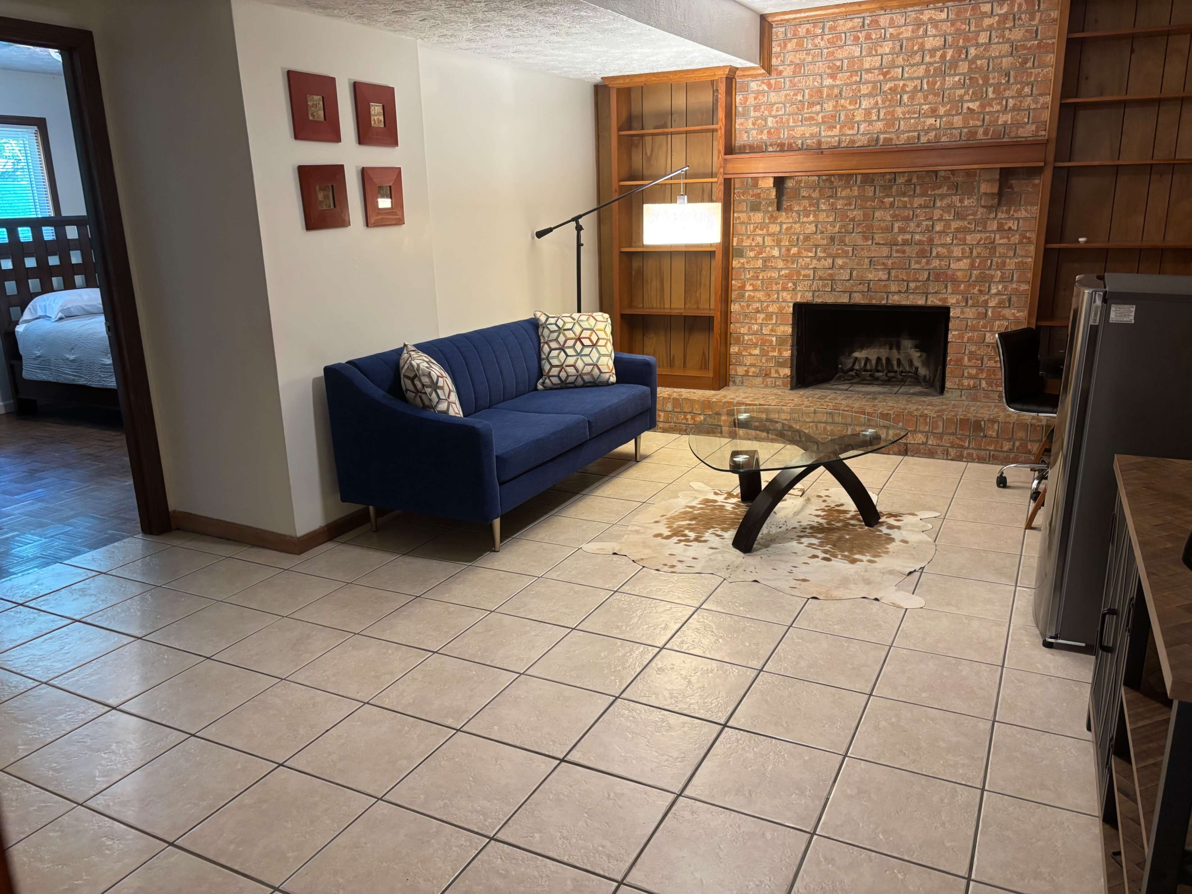 The image shows a living room with a blue couch, a coffee table, and a brick fireplace, featuring a cowhide rug on the tiled floor.