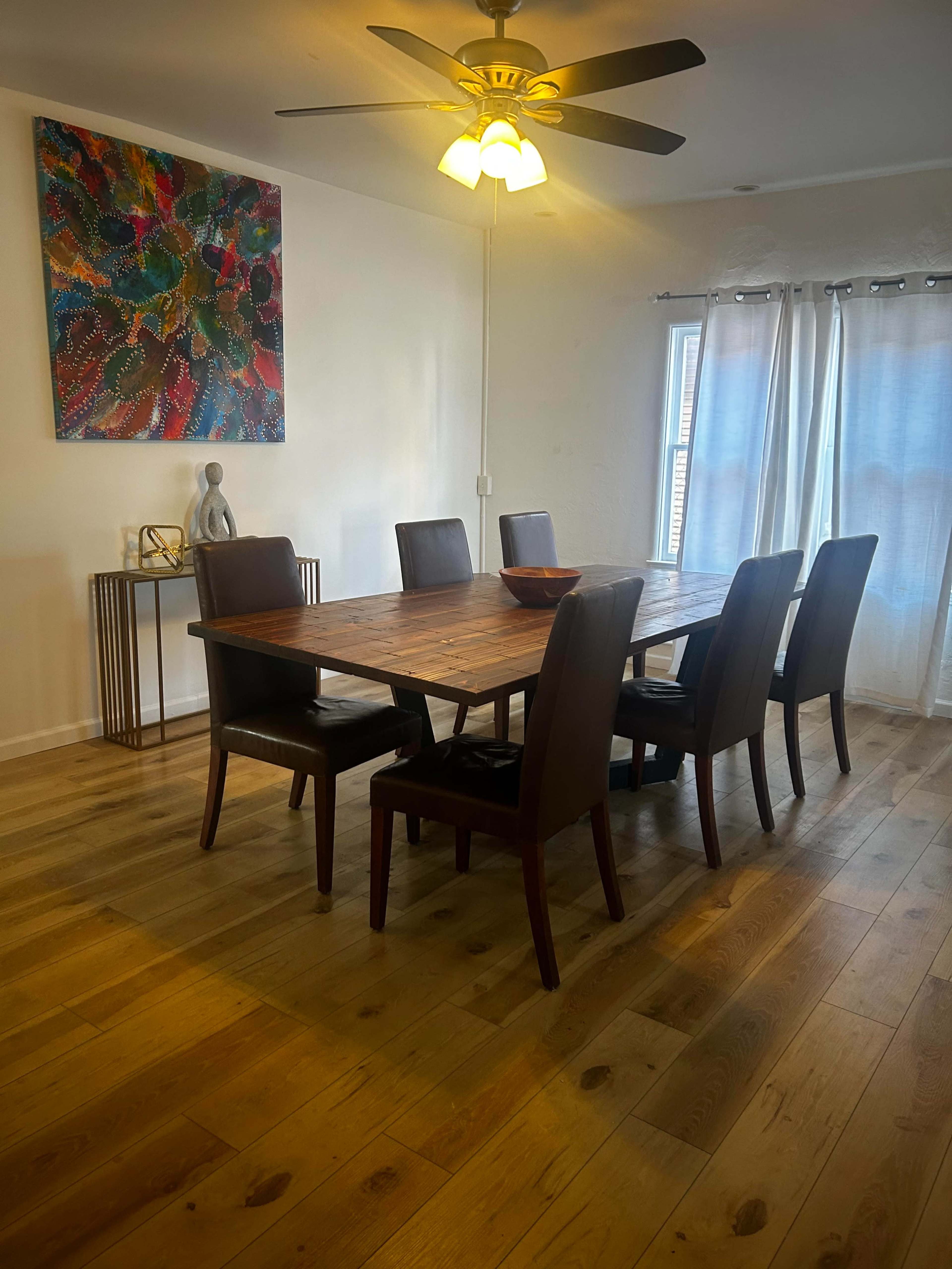 A wooden dining table surrounded by six chairs is set in a bright room with a ceiling fan, artwork on the wall, and large windows covered by sheer curtains.