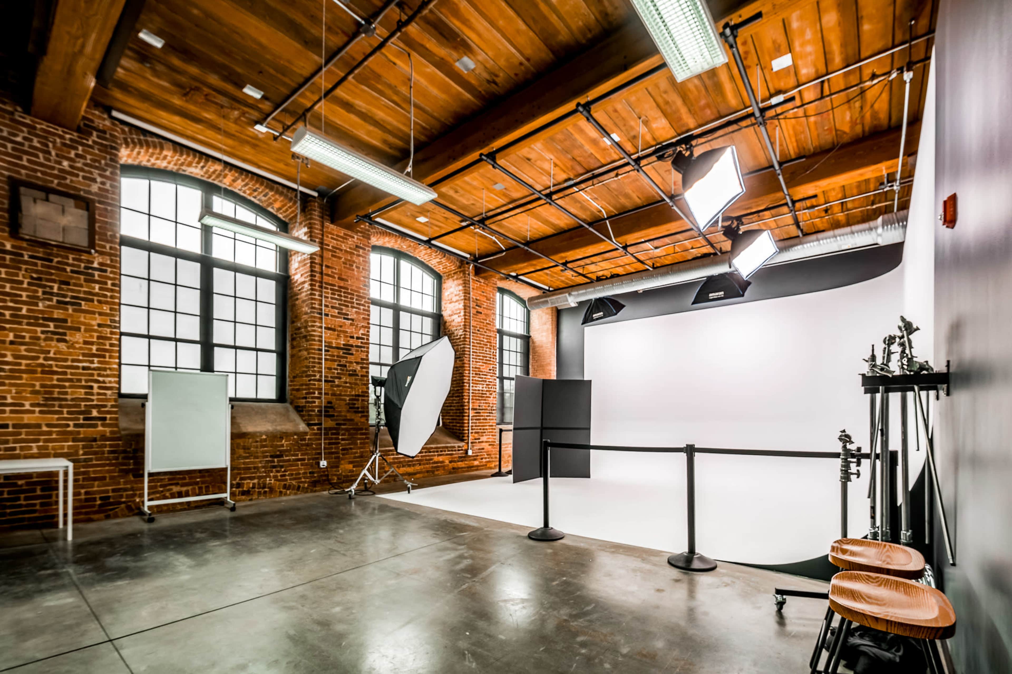 The image shows a photography studio with exposed brick walls, large windows, and professional lighting equipment arranged around a blank white backdrop.
